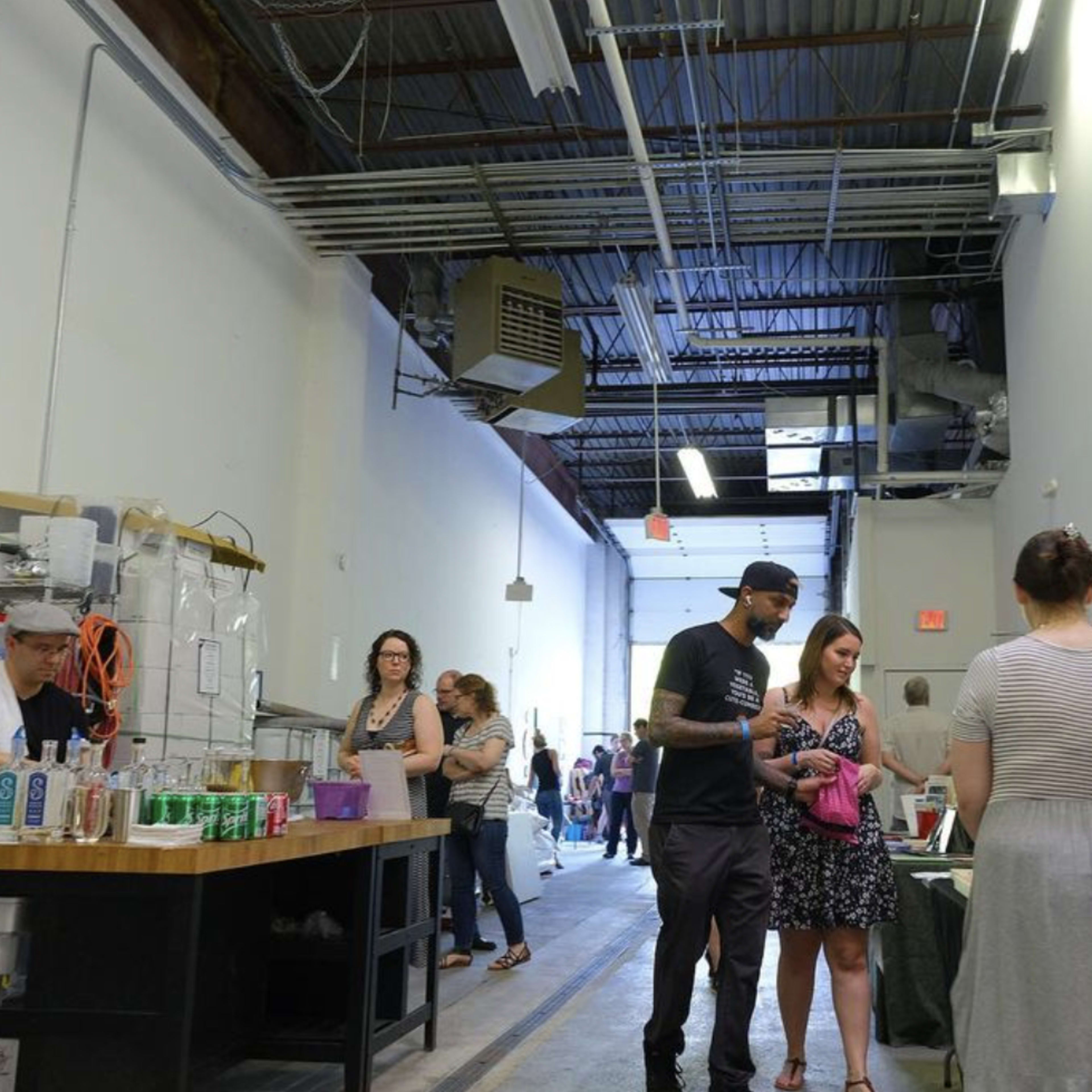 A group of people engages in conversation and browsing along an open corridor, with tables displaying various items and a counter serving beverages on one side.