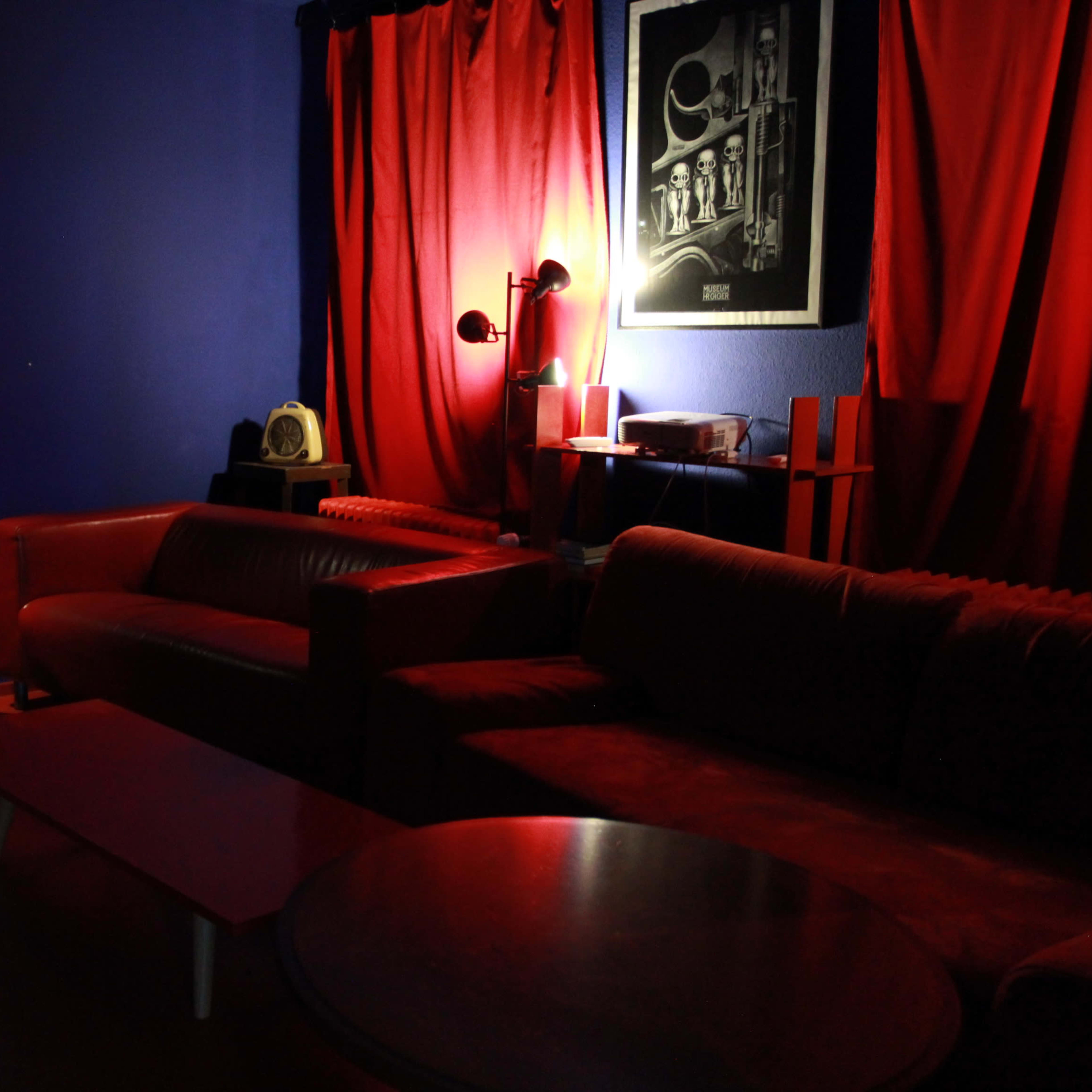 A dimly lit room with red curtains and a red sofa, featuring tables and a wall-mounted artwork.