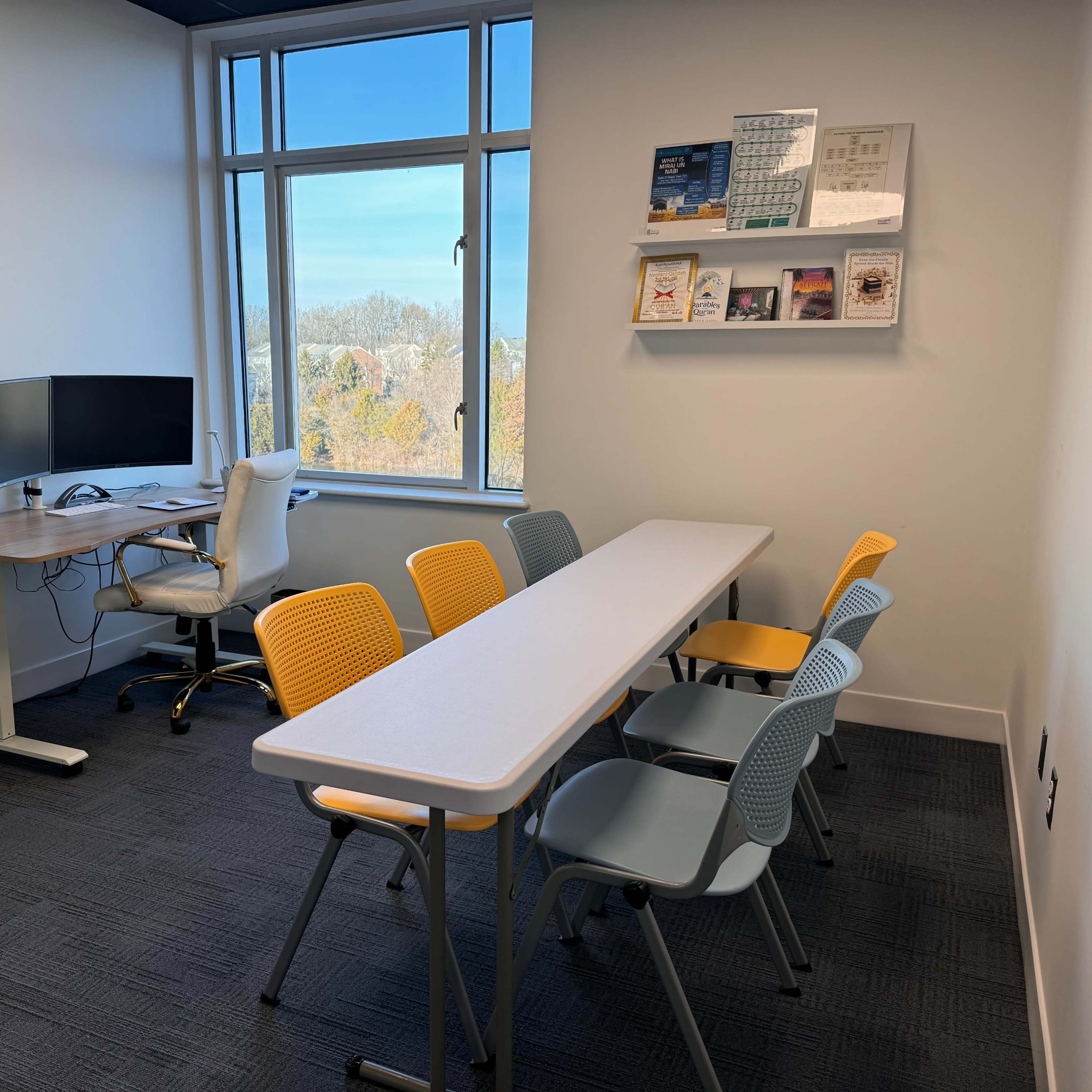 A small meeting room with a long table surrounded by chairs, a desk with a computer, and a window providing outside views.