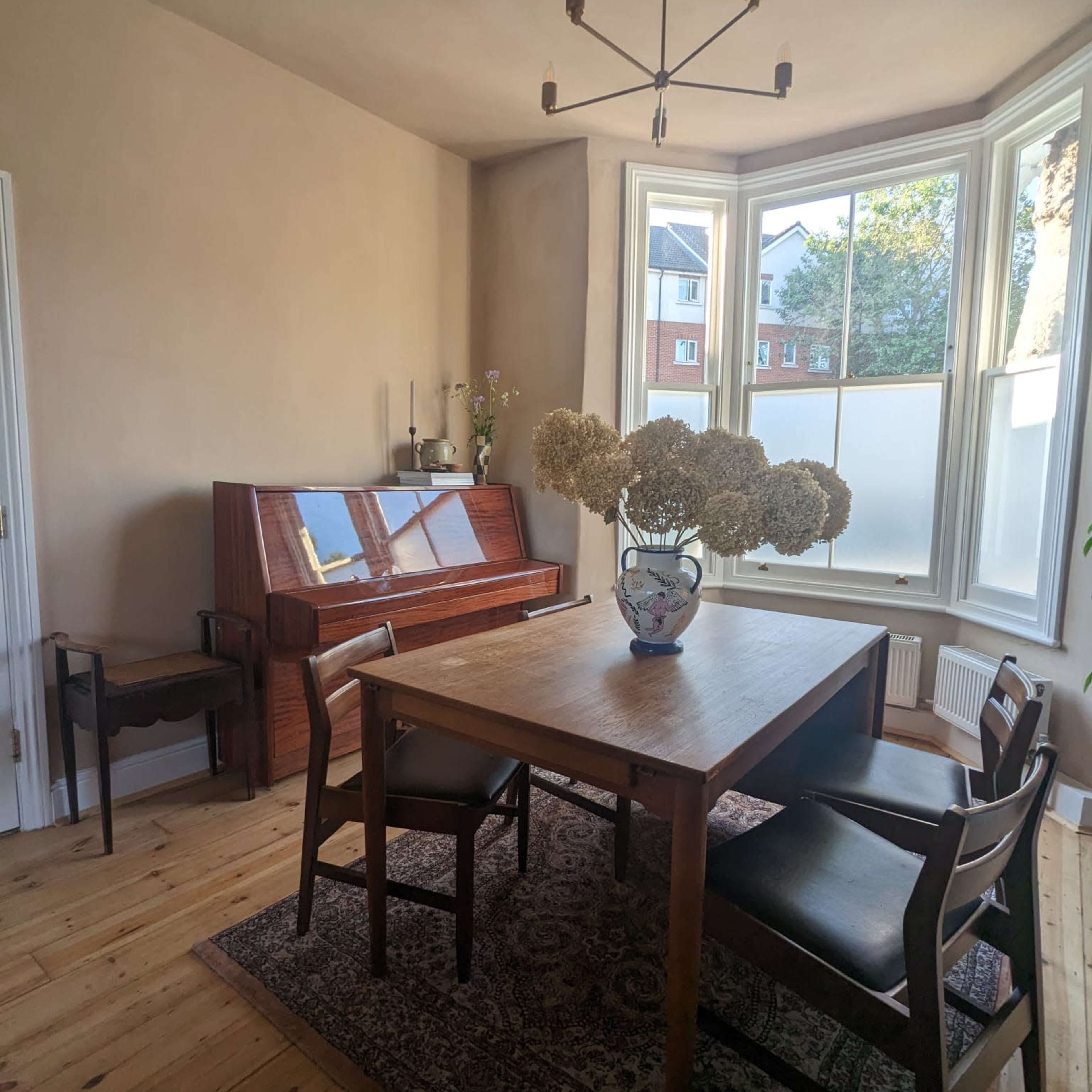A simple dining area features a wooden table surrounded by chairs, a piano against the wall, and a vase of dried flowers on the table.