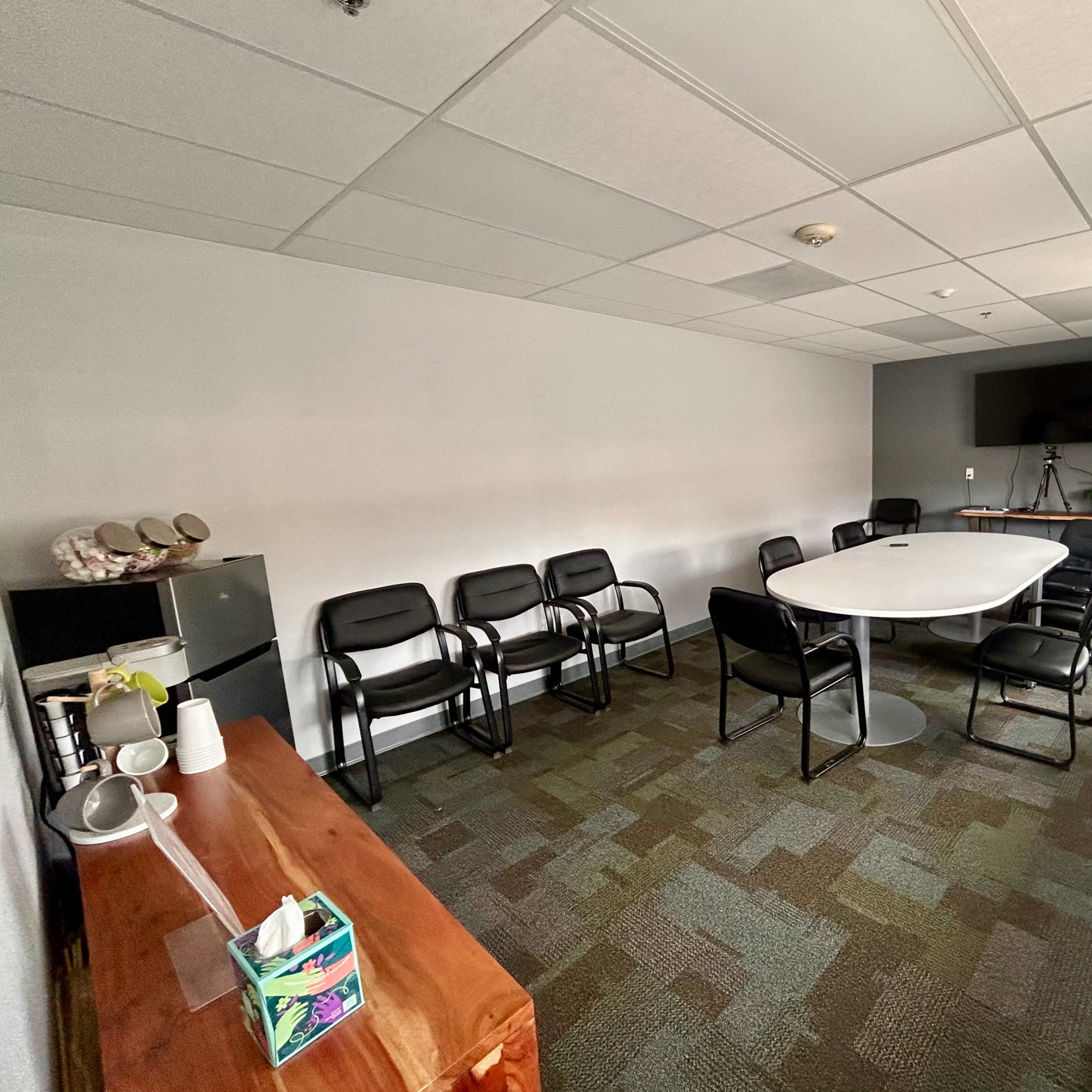 A conference room with a long oval table surrounded by black chairs, a television mounted on the wall, and a whiteboard in the corner.