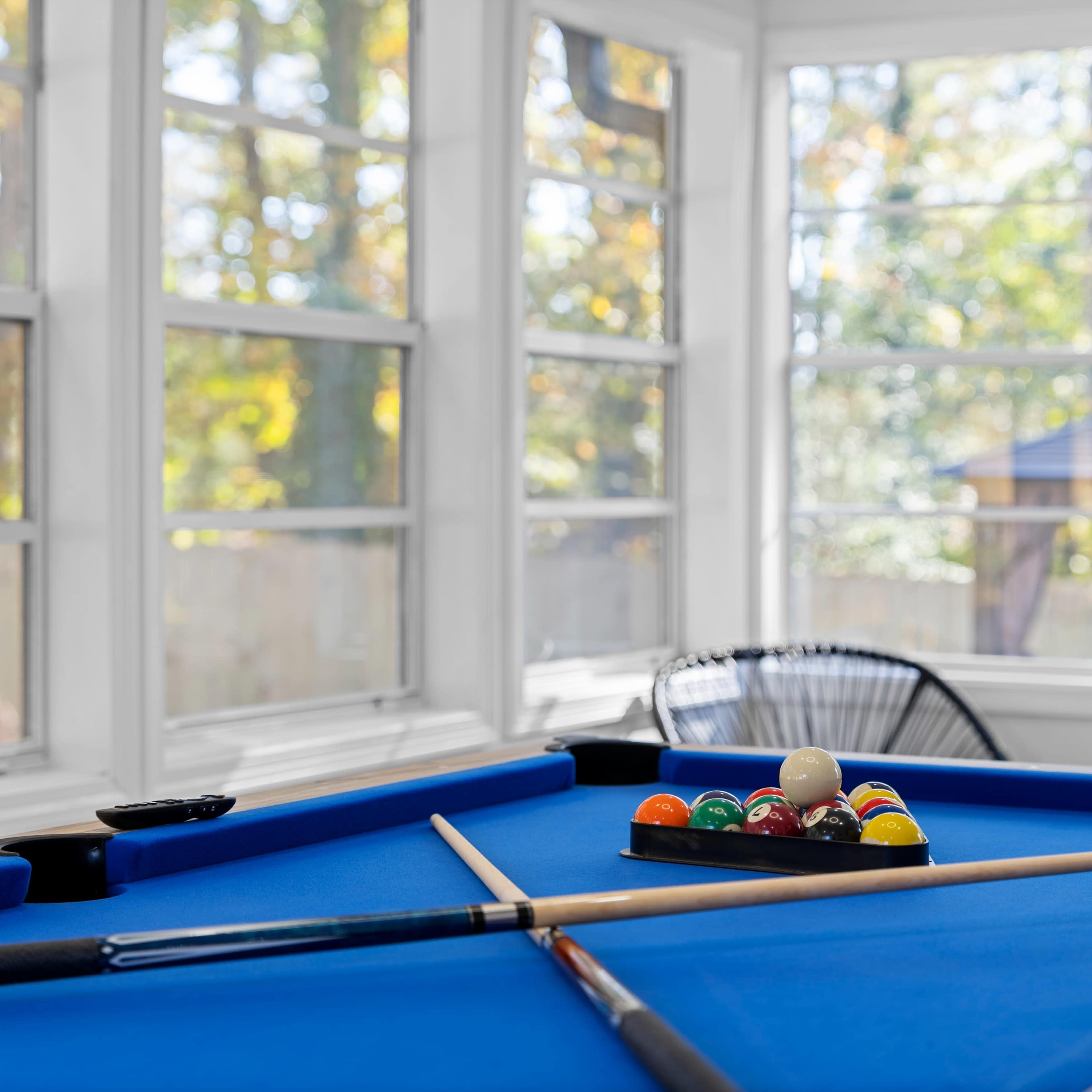 A blue pool table with cue sticks and a rack of billiard balls is set up in a sunlit room surrounded by large windows.
