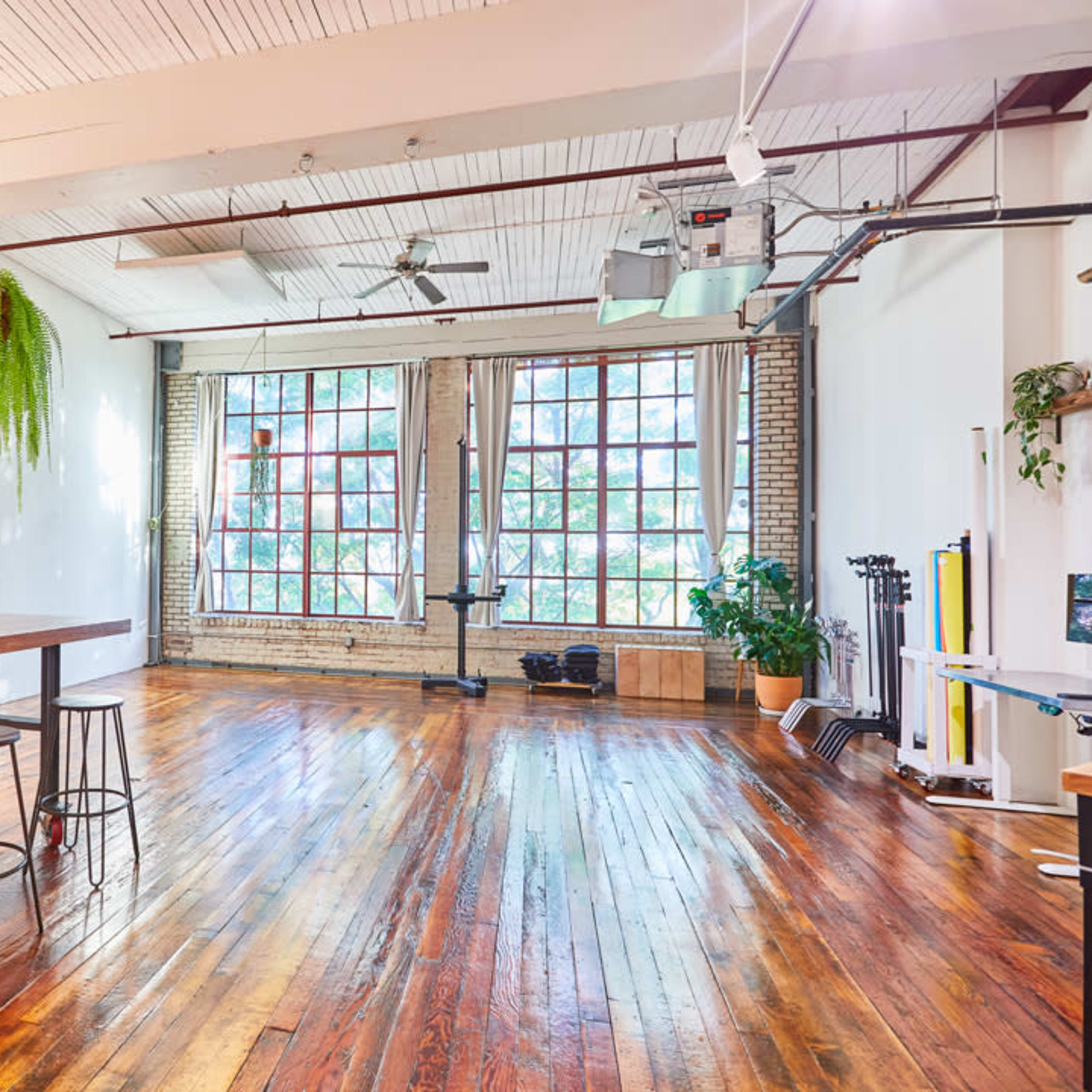 A spacious, well-lit interior with hardwood floors, a kitchen area featuring wooden cabinets and appliances, large windows, and a hanging plant.