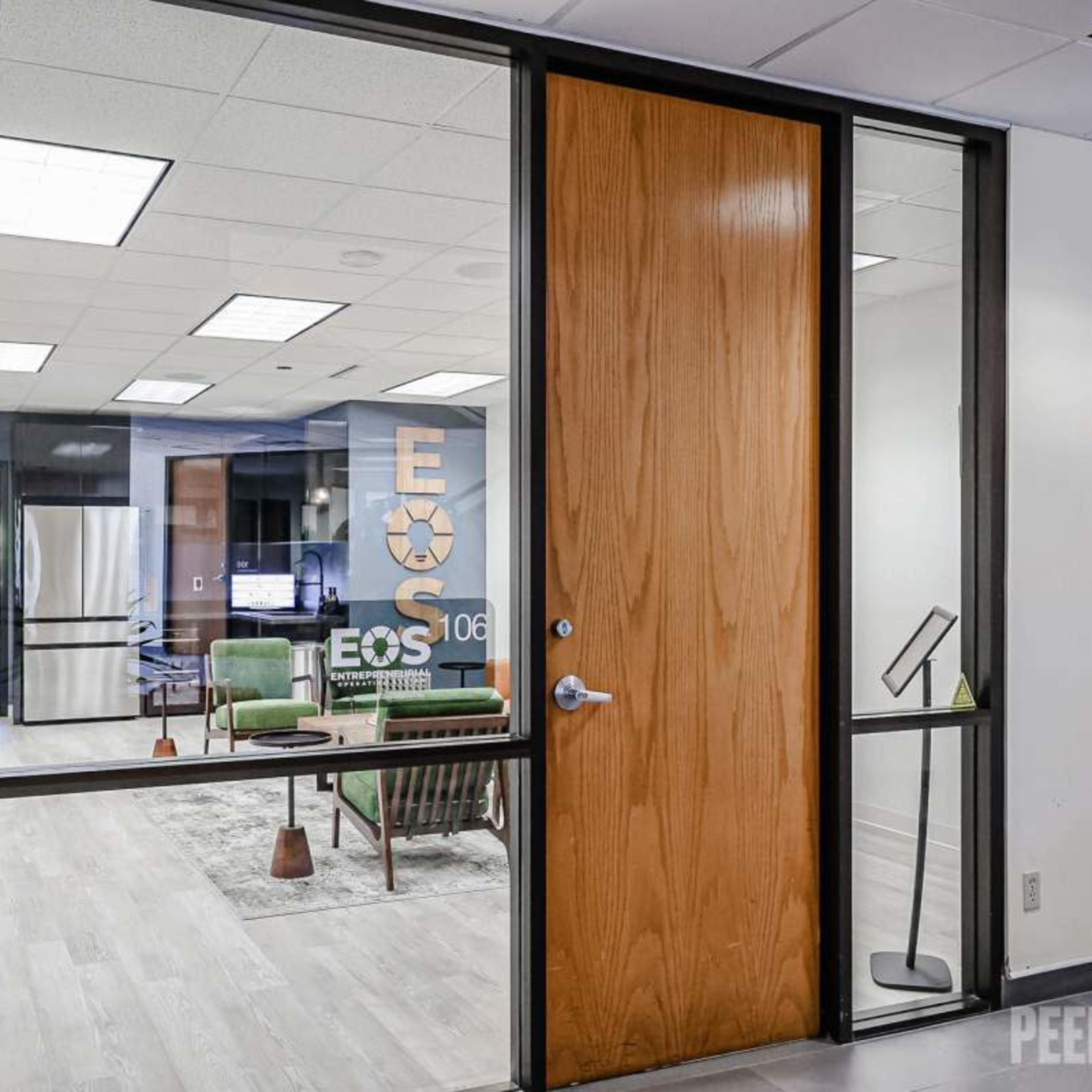 A modern office space visible through a glass door, featuring a sitting area with green furniture and an open layout.