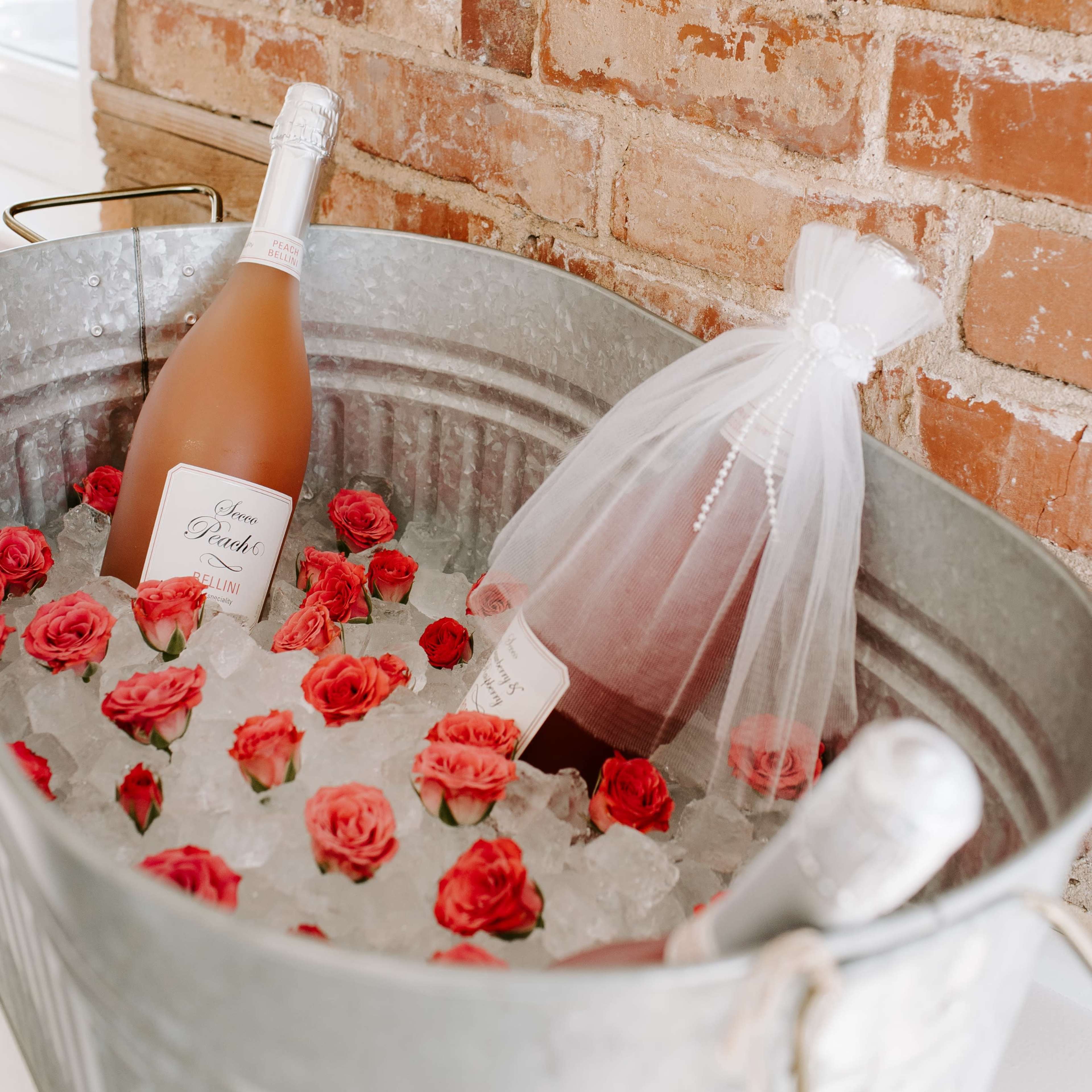 A galvanized metal tub filled with ice, bottles of rosé wine, and red rose petals sits against a brick wall.