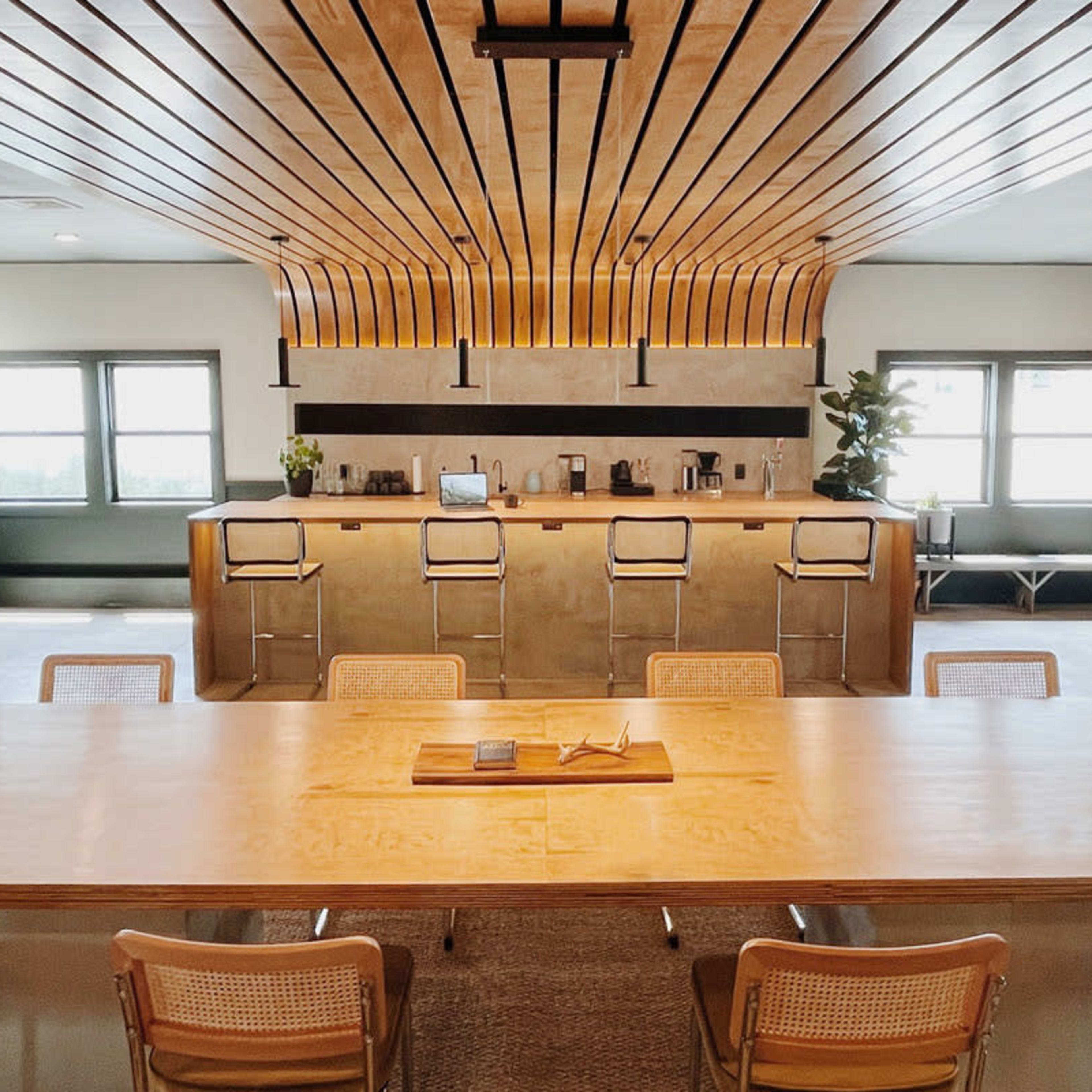 A modern office breakroom featuring a long wooden table, several chairs, and a kitchen area with bar stools, cabinets, and coffee machines.