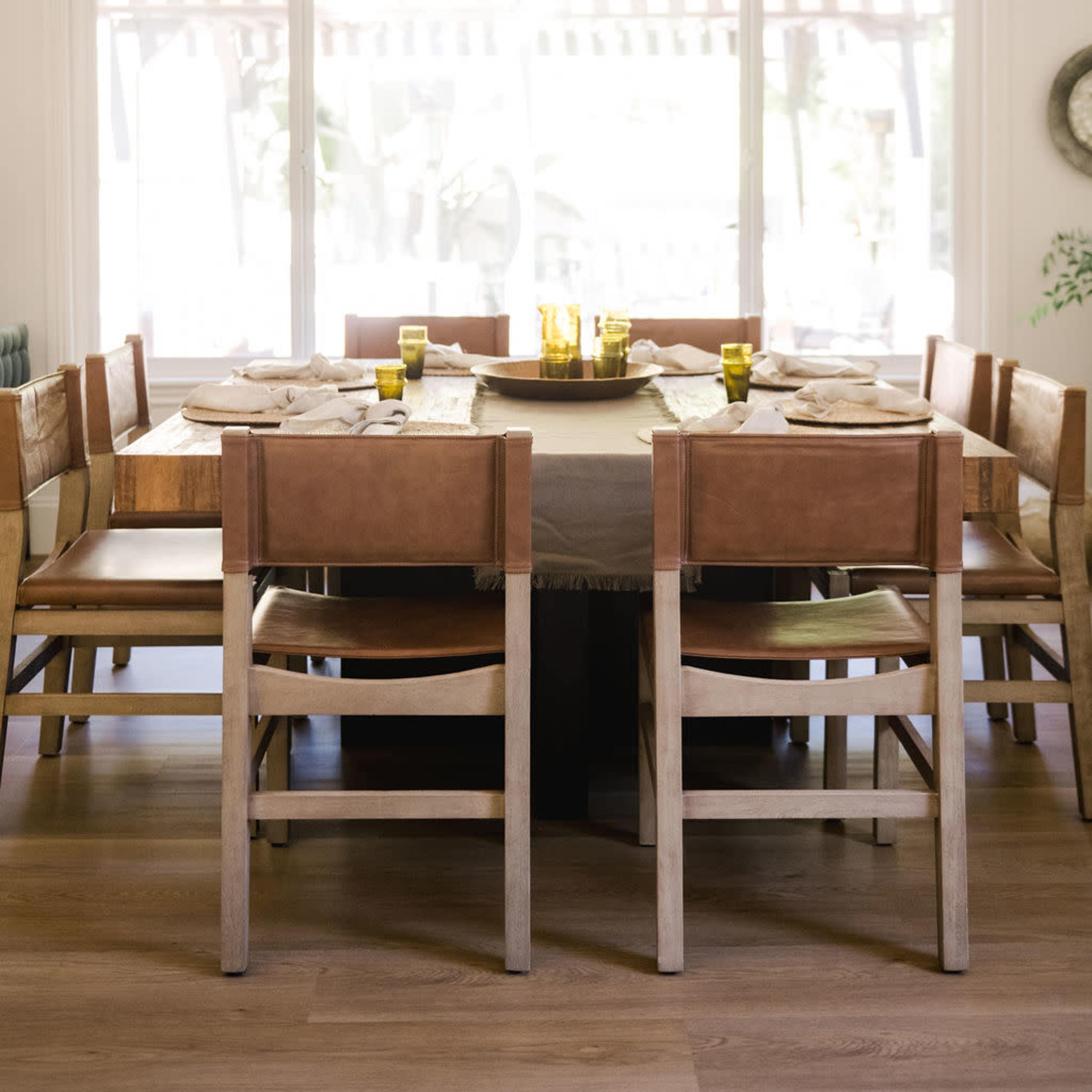A large wooden dining table surrounded by leather and wooden chairs is set for a meal, with green glasses and plates arranged around it in a well-lit room.