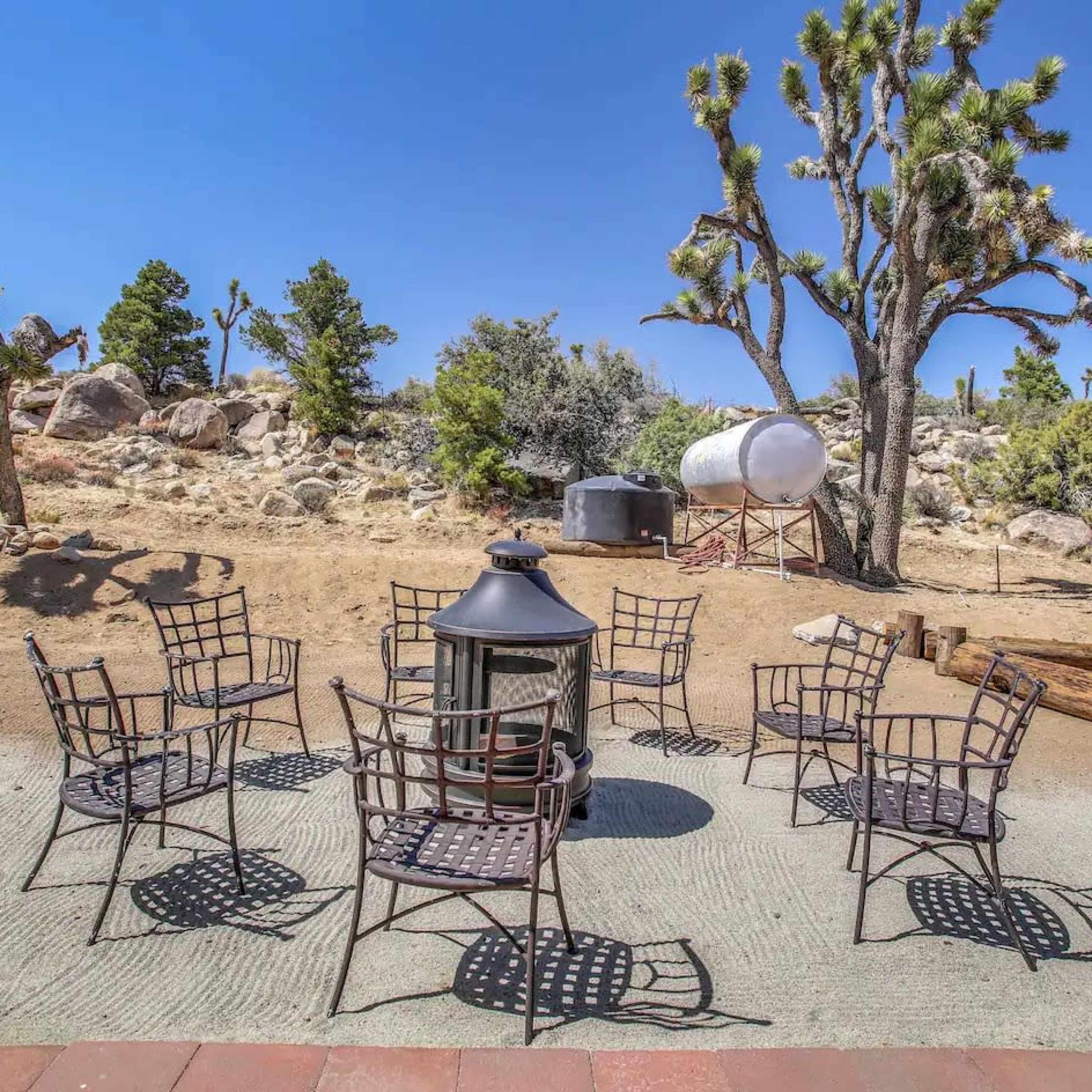 A circle of metal chairs surrounds a lantern on a stone patio, with desert vegetation and rocky terrain in the background.