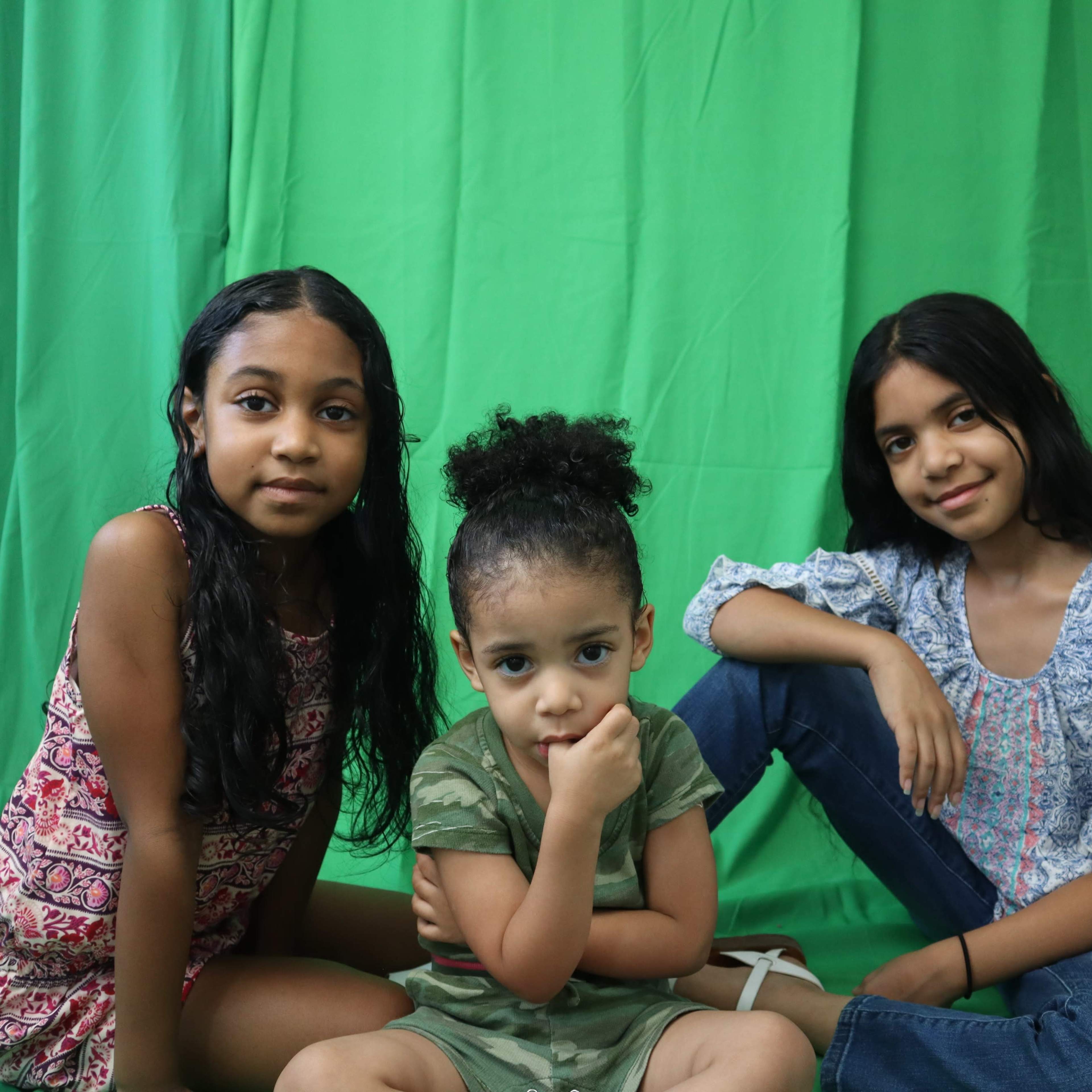Three young girls pose in front of a green backdrop, with one sitting in the center and the others on either side.