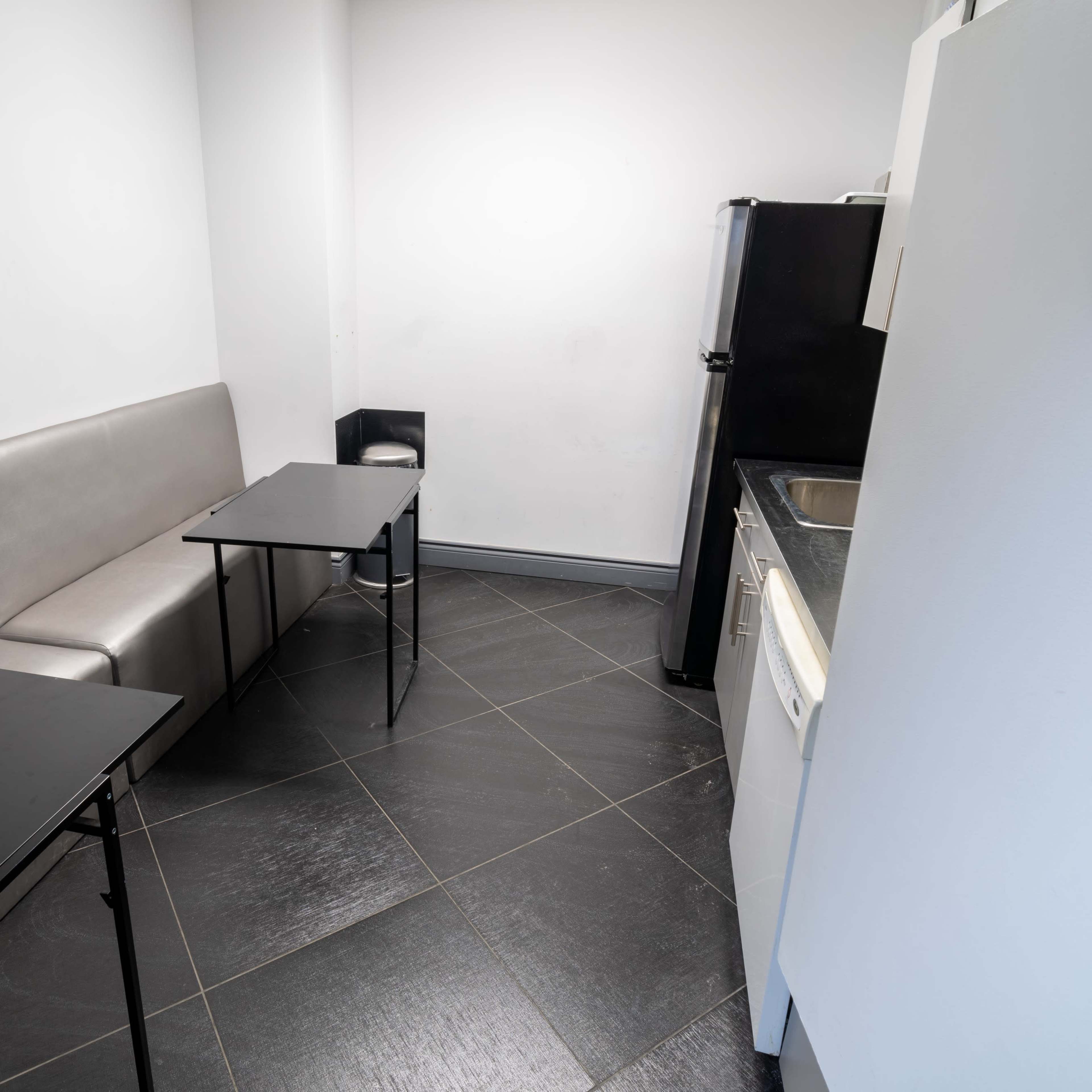 A small kitchen area features a gray bench seating and two black tables, with a refrigerator and sink visible in the background.