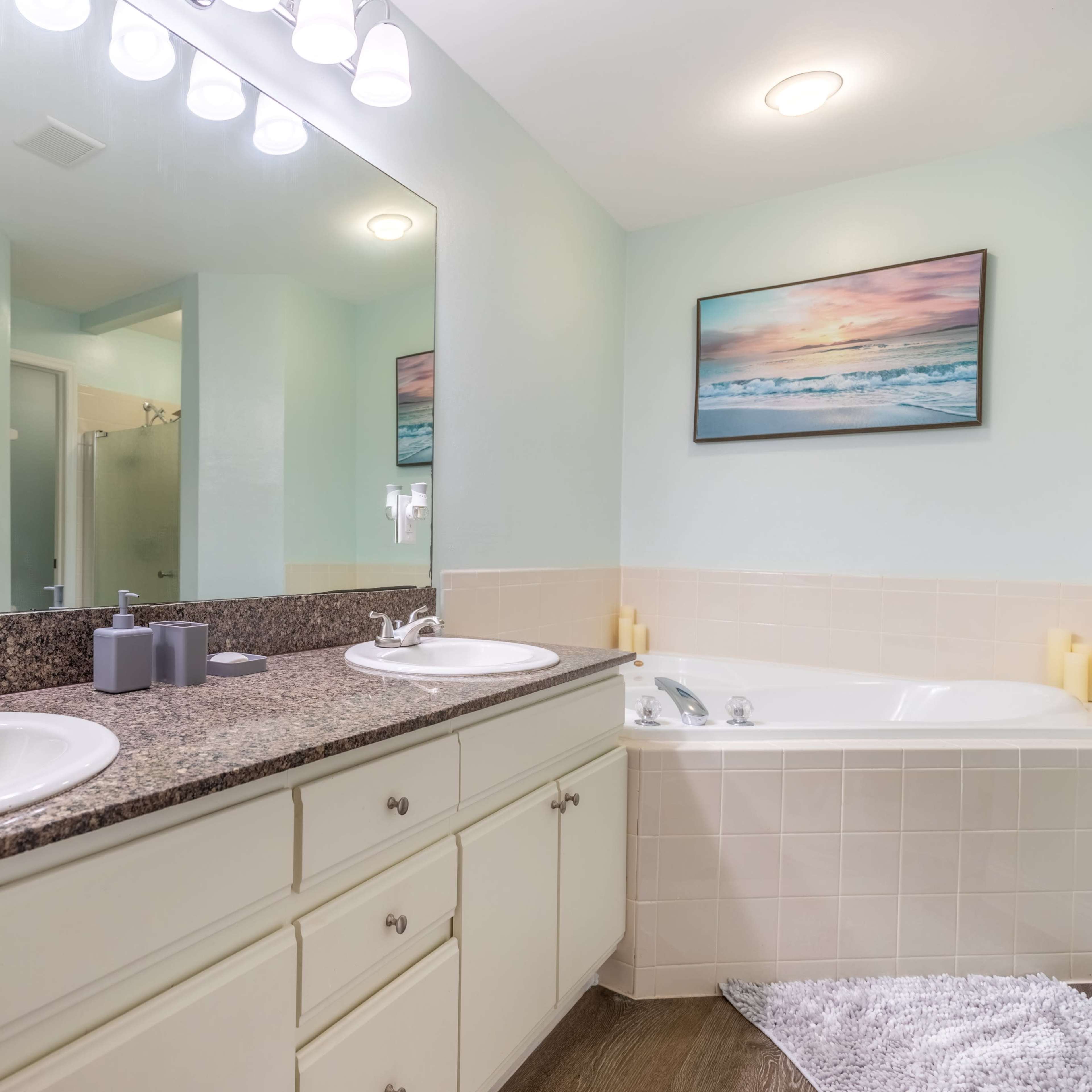 A bathroom with double sinks, a bathtub, and a mirrored vanity, set against light blue walls and a beach-themed painting.