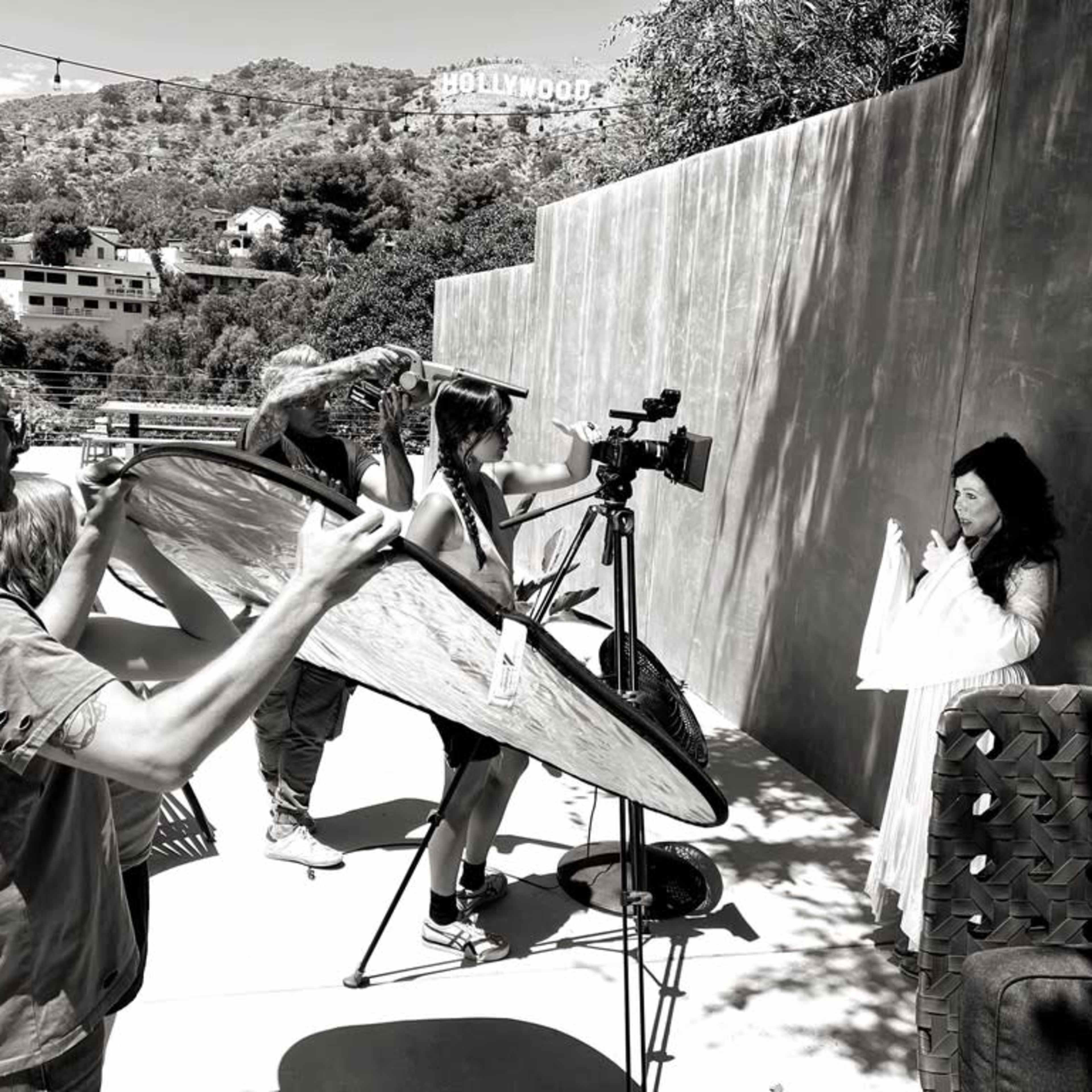 A film crew is set up outdoors with a camera and lighting equipment, preparing to shoot a scene featuring a woman in a white dress against a textured wall, with the Hollywood sign visible in the background.