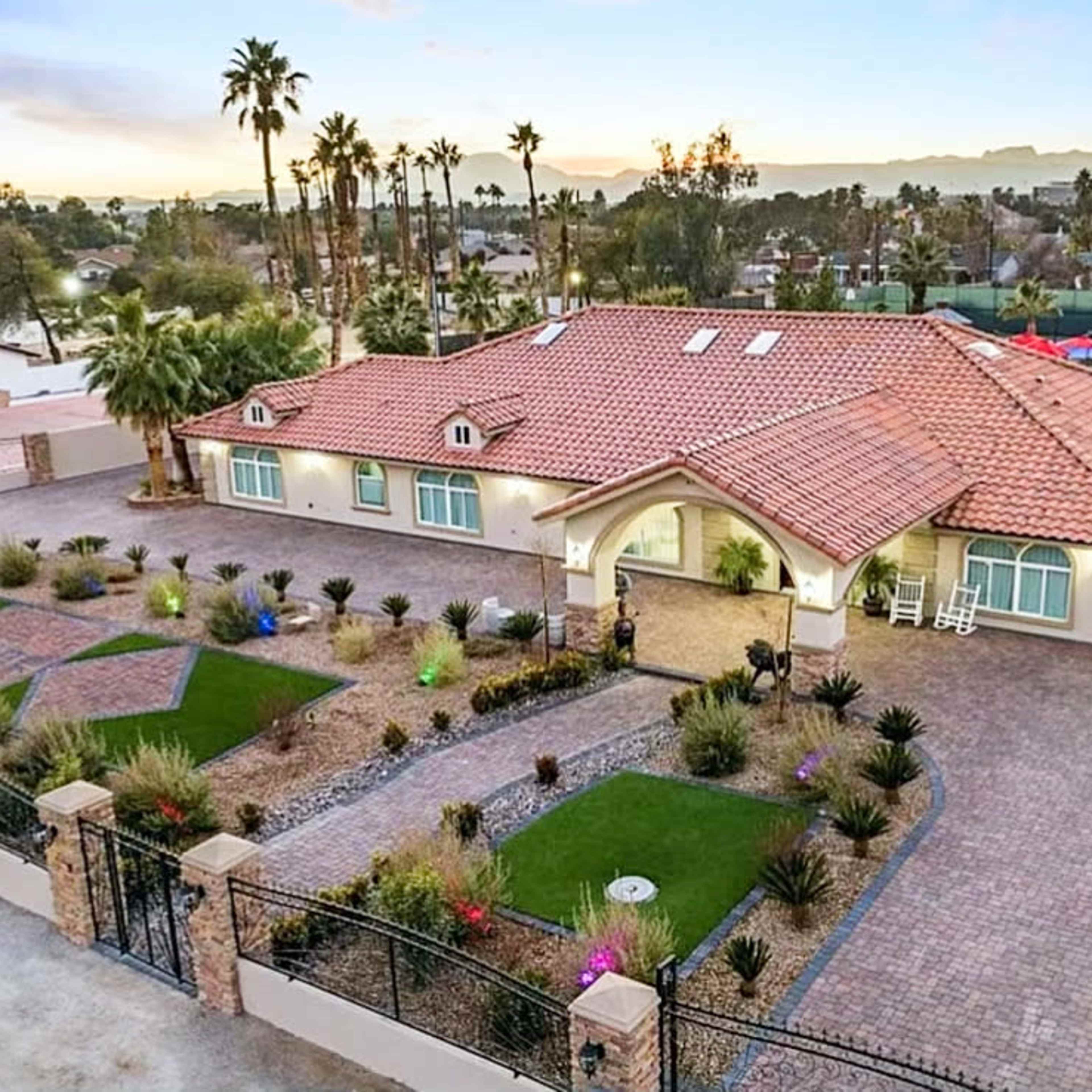 A large, single-story home with a red-tiled roof, surrounded by landscaped gardens and palm trees in a spacious driveway.
