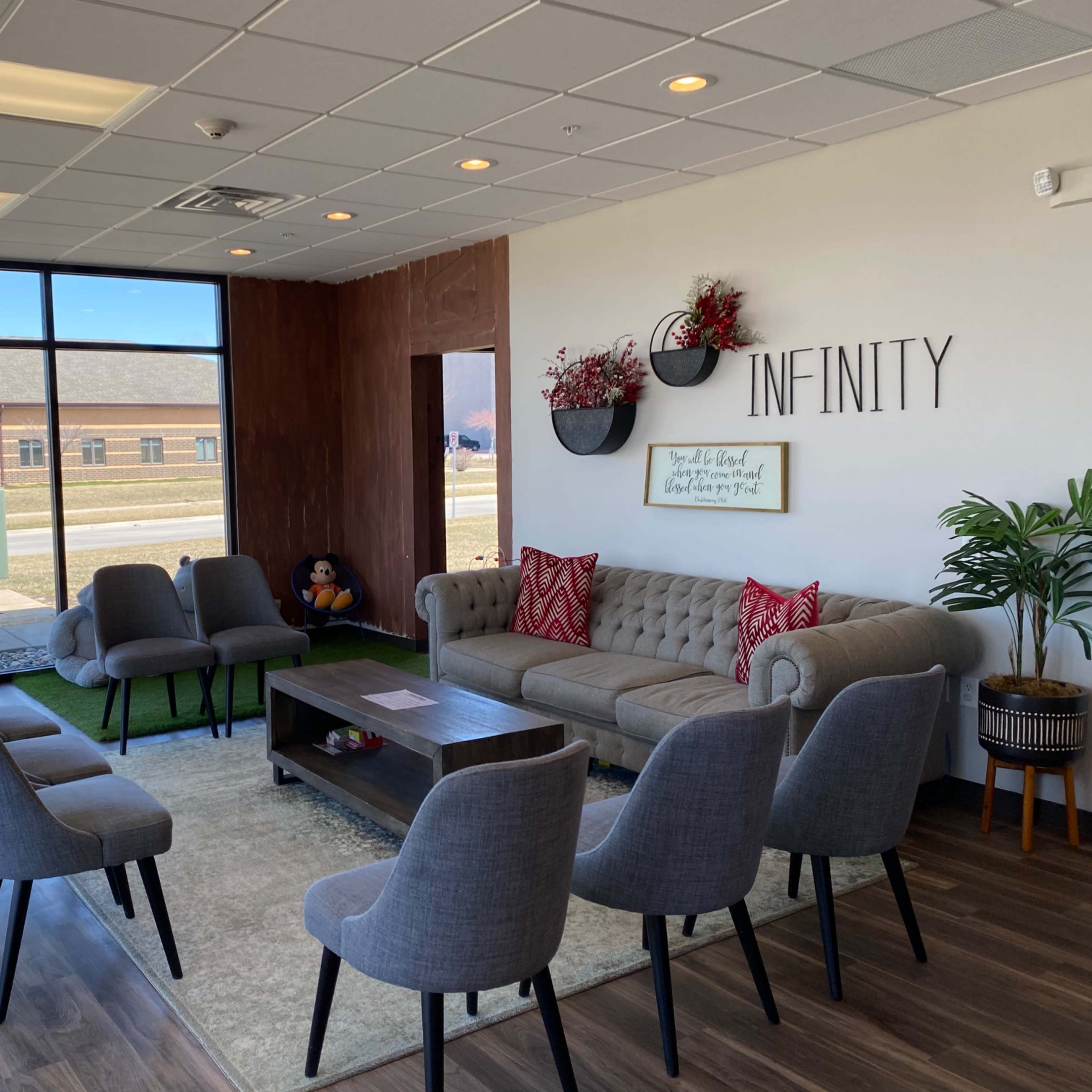 A modern waiting area with a beige sofa, a coffee table, and multiple gray chairs arranged around the room, featuring large windows that provide natural light.