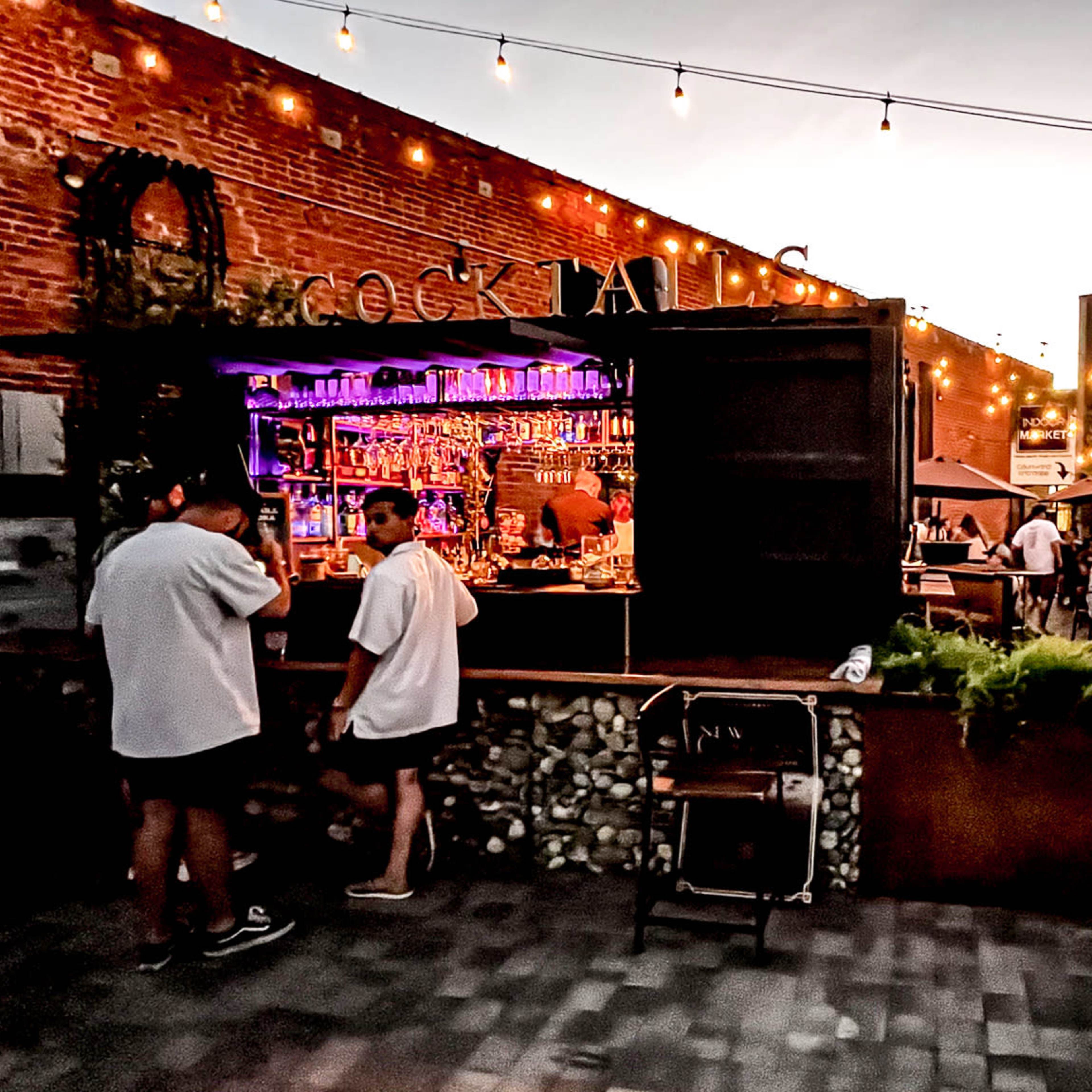 An outdoor bar scene features patrons interacting at a stylish bar illuminated by string lights, with people seated at tables nearby.