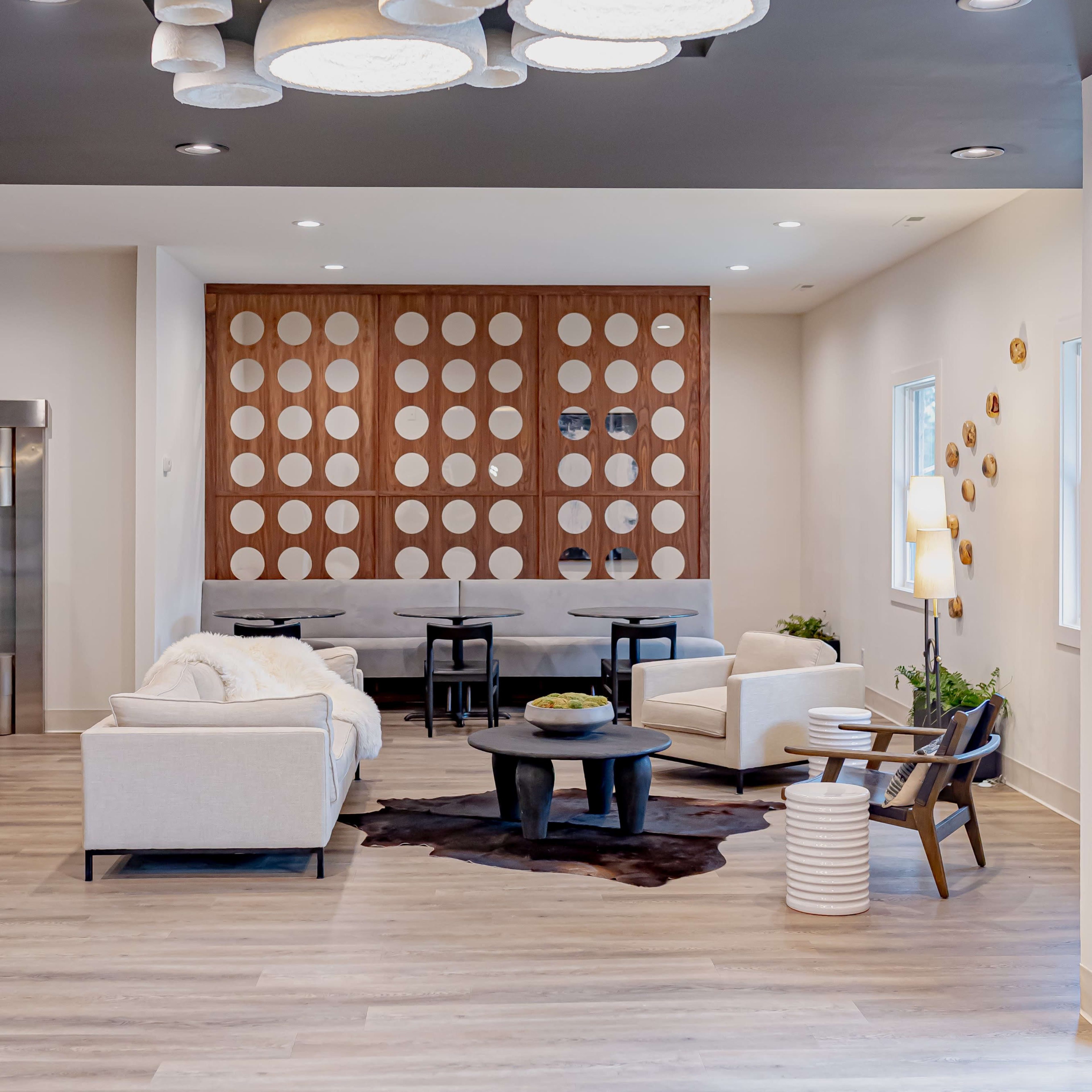 A modern lounge area with a minimalist design, featuring light-colored furniture, decorative wall panels, and a cluster of pendant lights overhead.