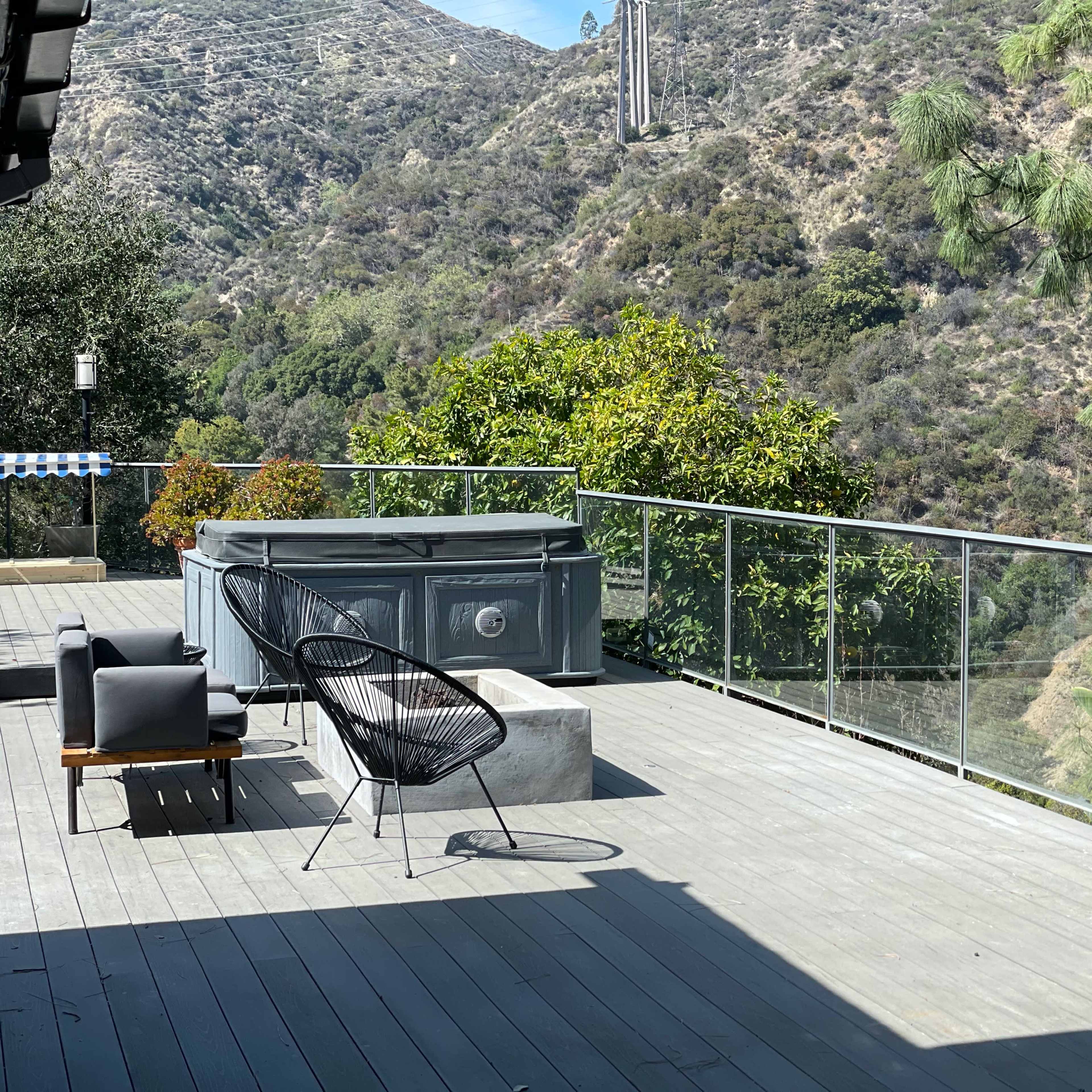 A deck overlooks a mountainous landscape, featuring a hot tub, an outdoor seating area, and greenery in the background.