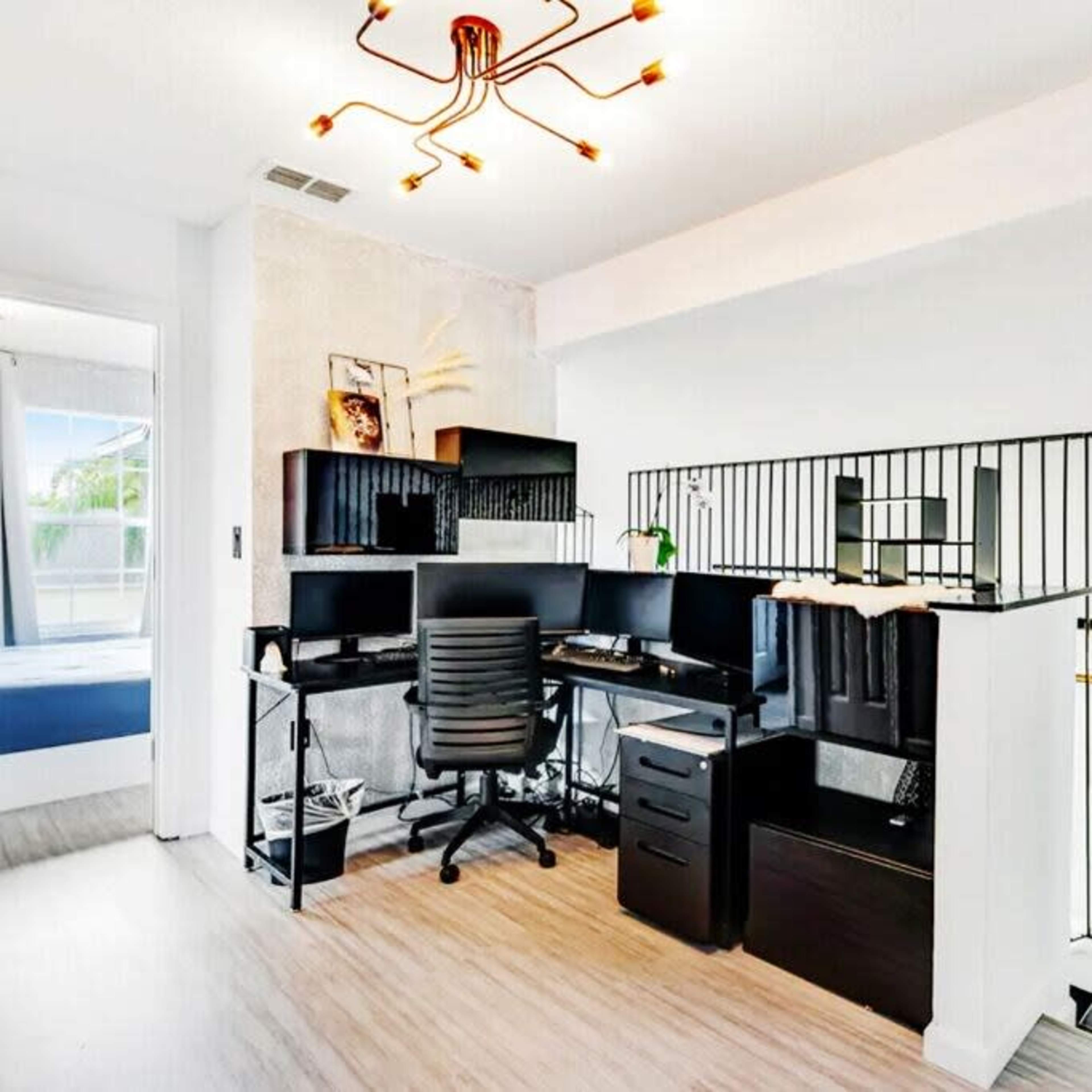 A modern workspace featuring a desk with two monitors, a black office chair, and a decorative overhead light, adjacent to a bedroom space.