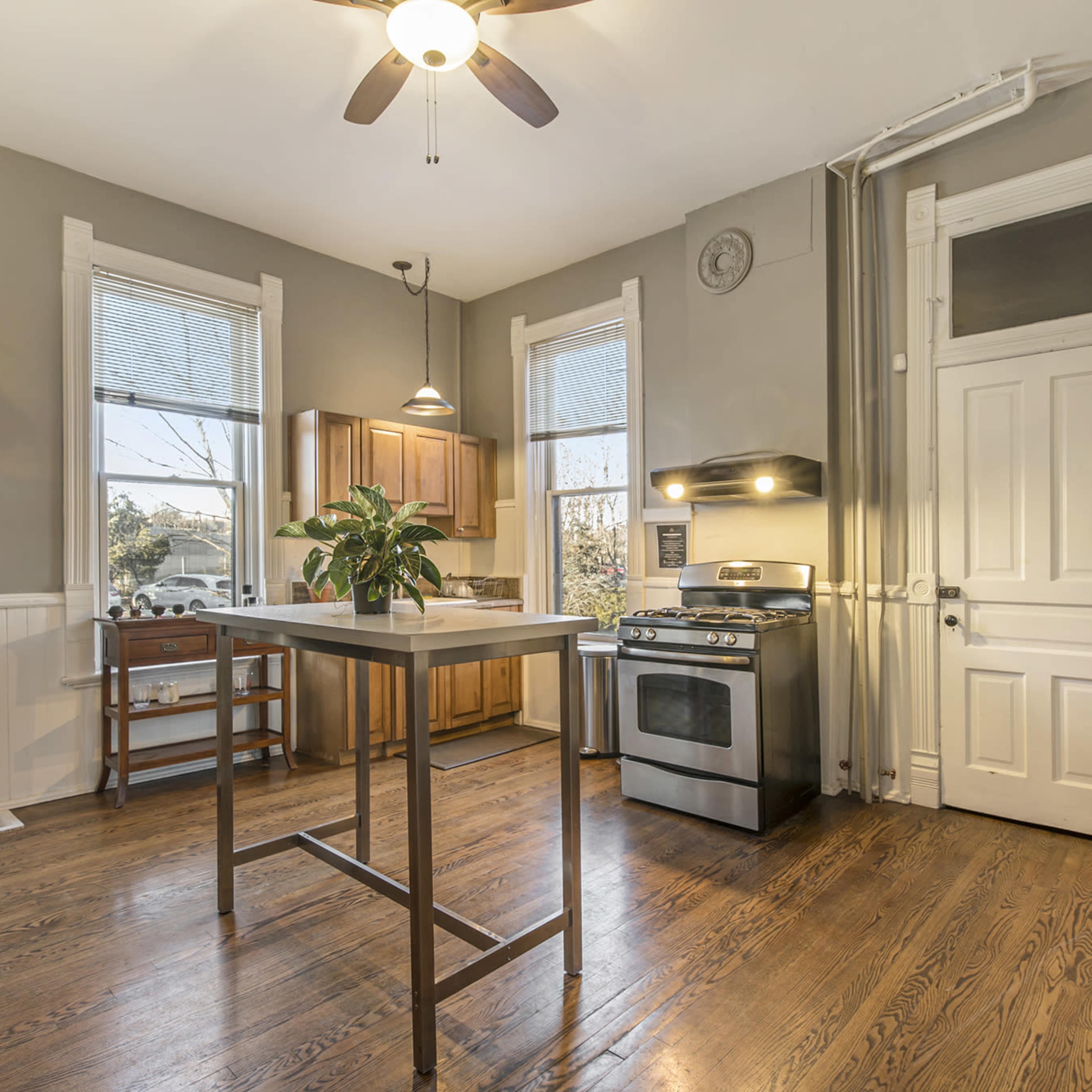 A kitchen with wooden floors, a stainless steel stove, and a central island with a plant on it, surrounded by cabinets and windows.