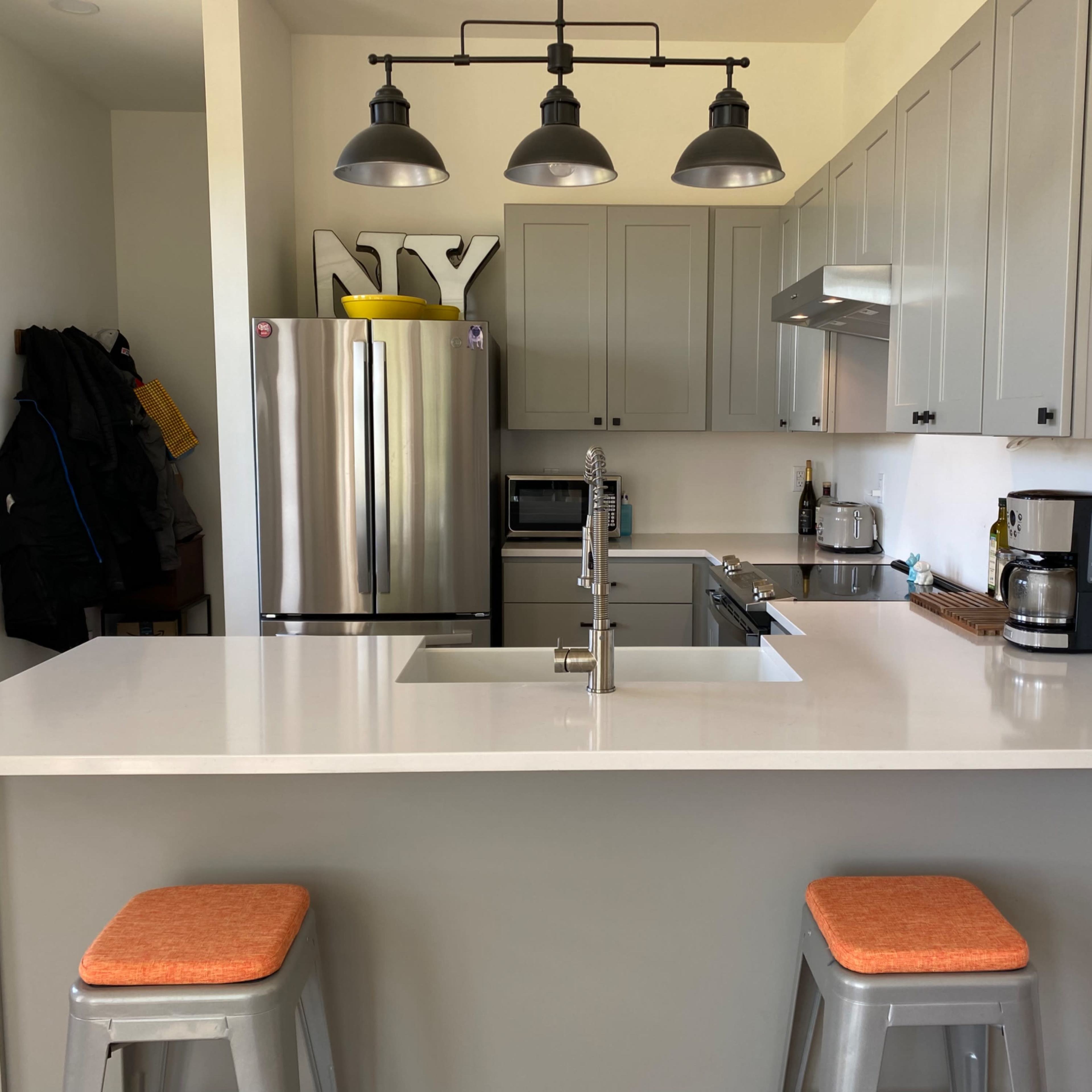 A modern kitchen with gray cabinets, stainless steel appliances, a large island with a white countertop, and two orange stools.