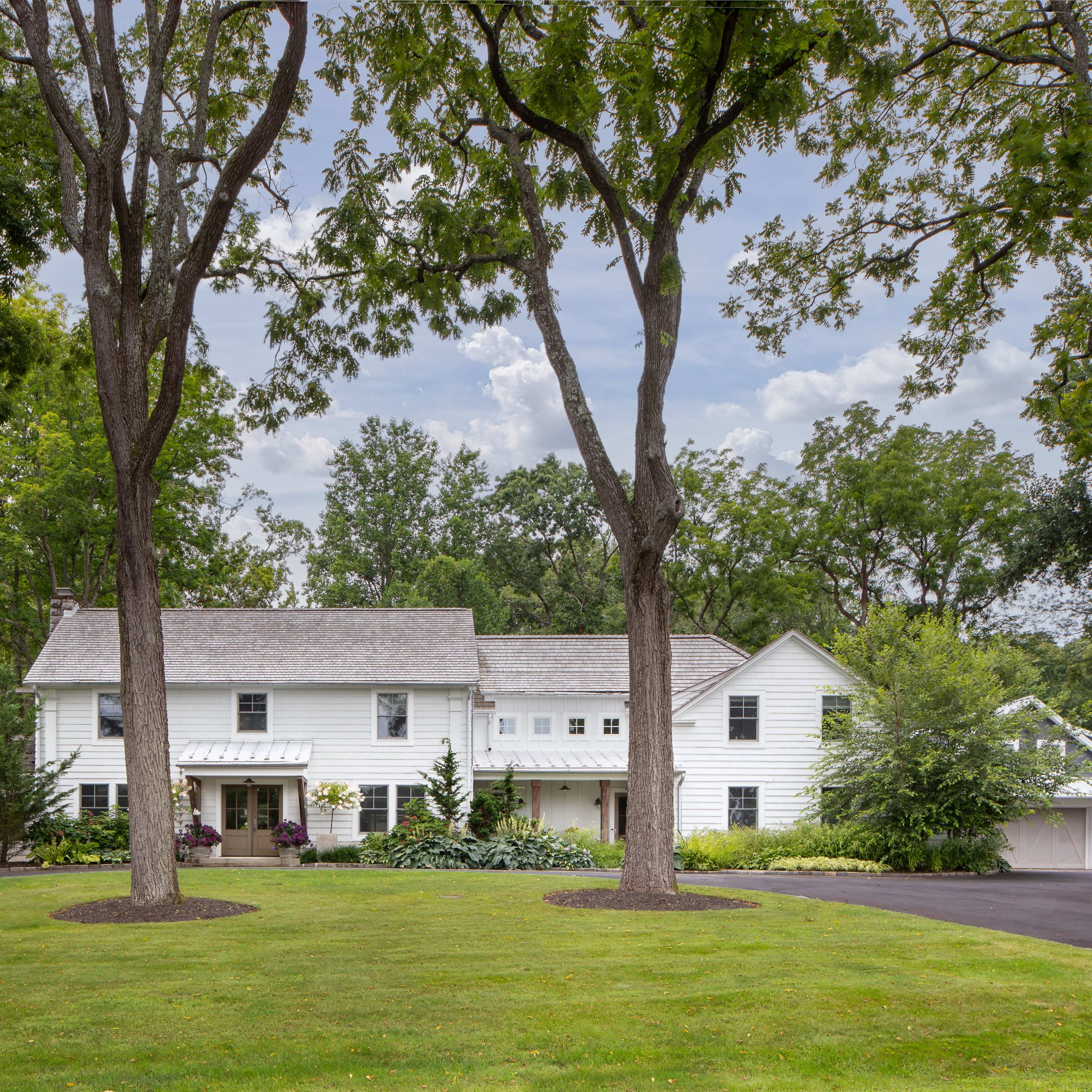 A large, white two-story house is situated among tall trees on a manicured lawn, with a paved driveway leading up to it.