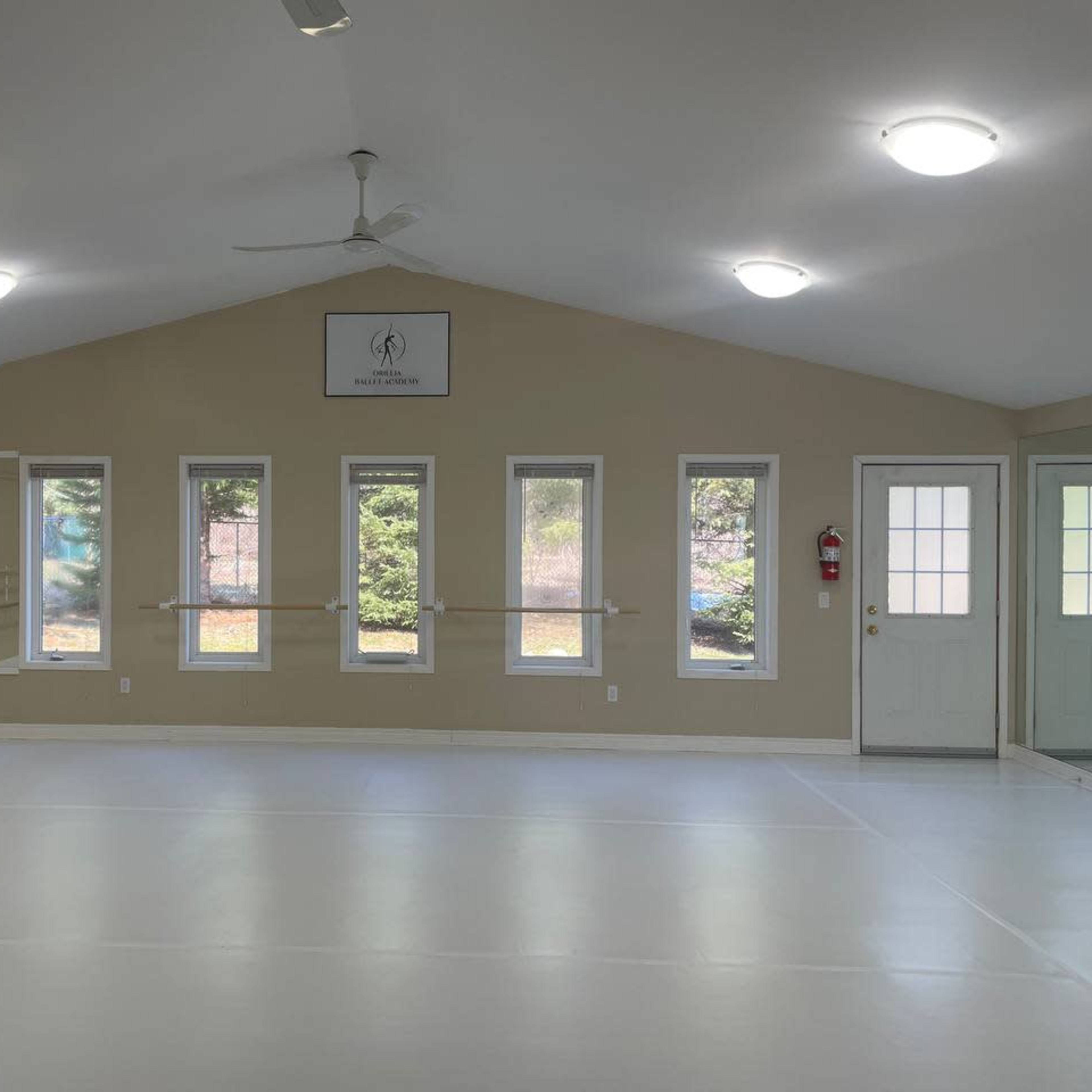 An empty dance studio with large windows, a mirror on one wall, and wooden ballet bars along another wall, illuminated by overhead lights.
