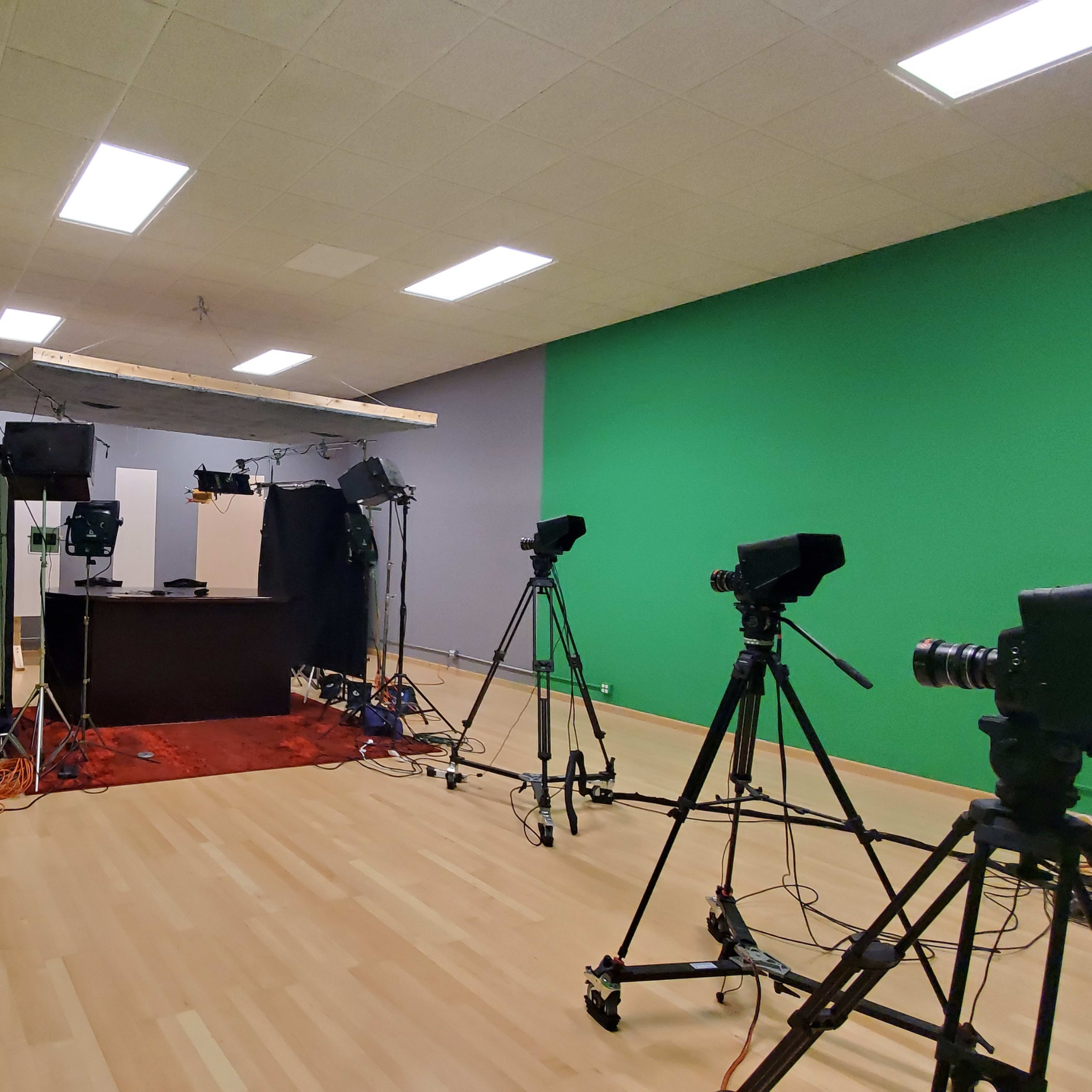 A video production studio with green backdrop walls, multiple cameras, and lighting equipment set up around a central workspace.