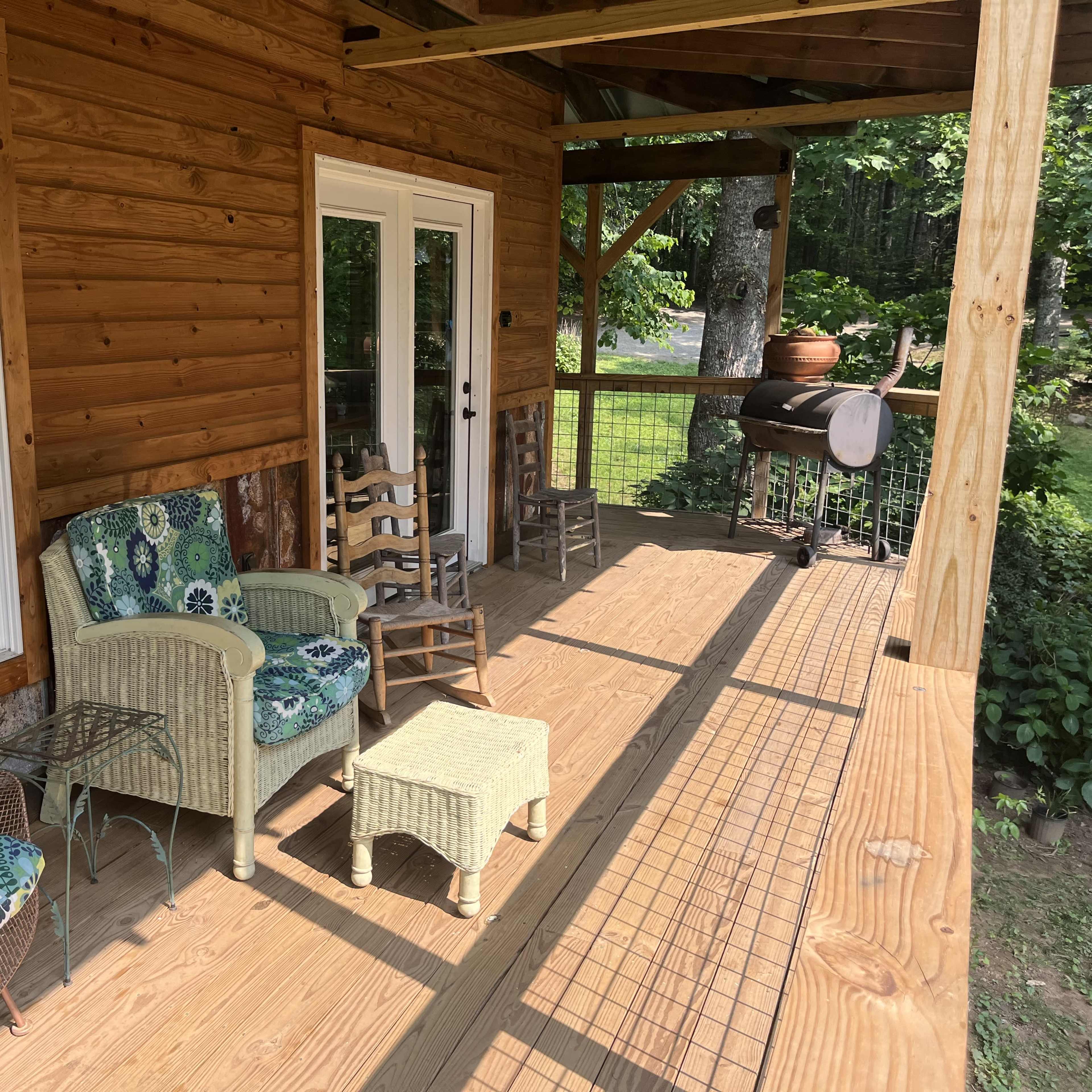 A wooden porch with wicker and metal chairs, a small table, and a grill, surrounded by greenery.
