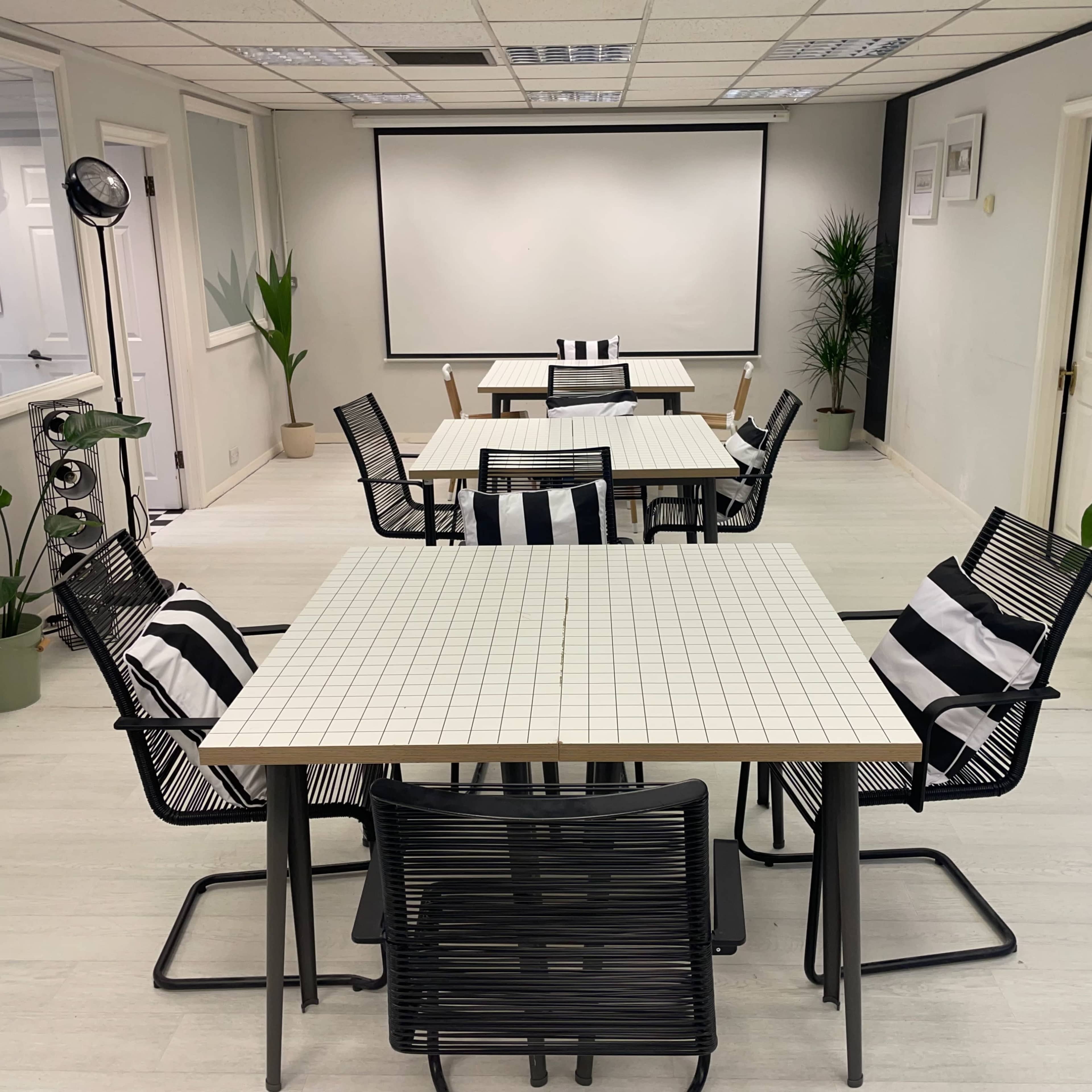 A modern meeting room features several square tables with striped chairs, surrounded by potted plants and a projector screen at the front.