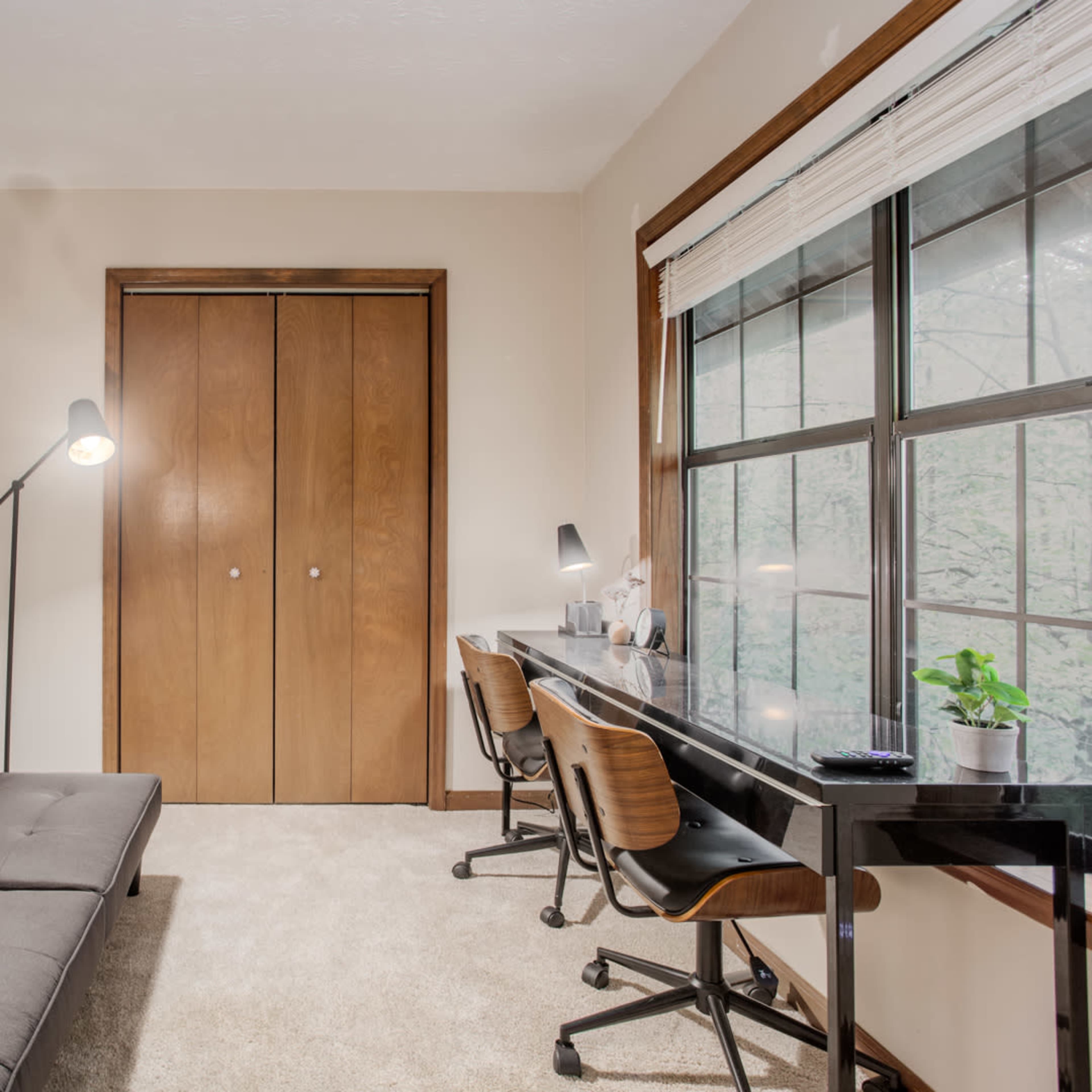 A small room features a gray sofa, a black desk with two chairs, and a large window overlooking greenery, with a framed picture of a cow on the wall.
