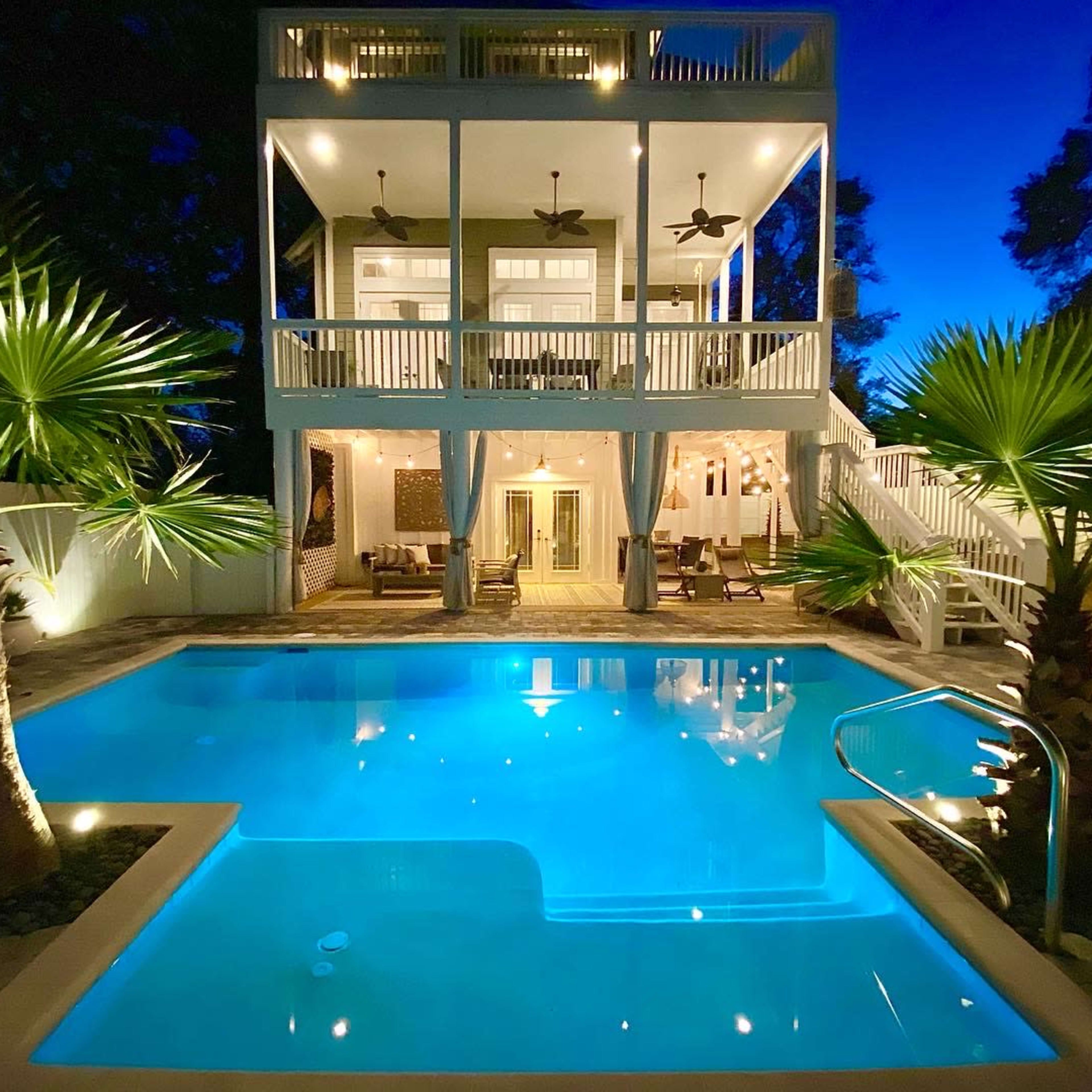 A two-story house with large balconies overlooks a illuminated swimming pool surrounded by palm trees at twilight.