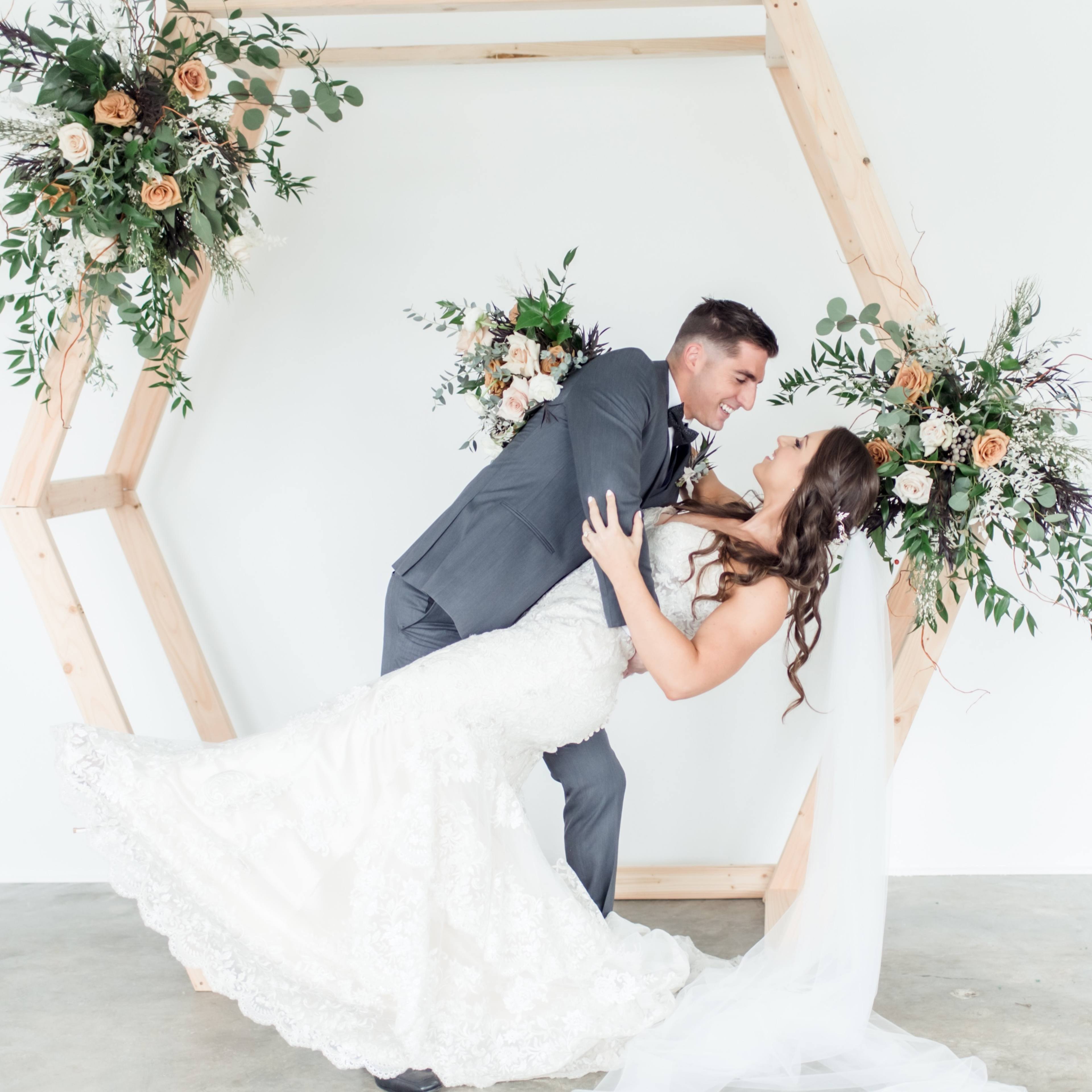 A couple poses in a dip under a wooden hexagonal arch decorated with flowers.