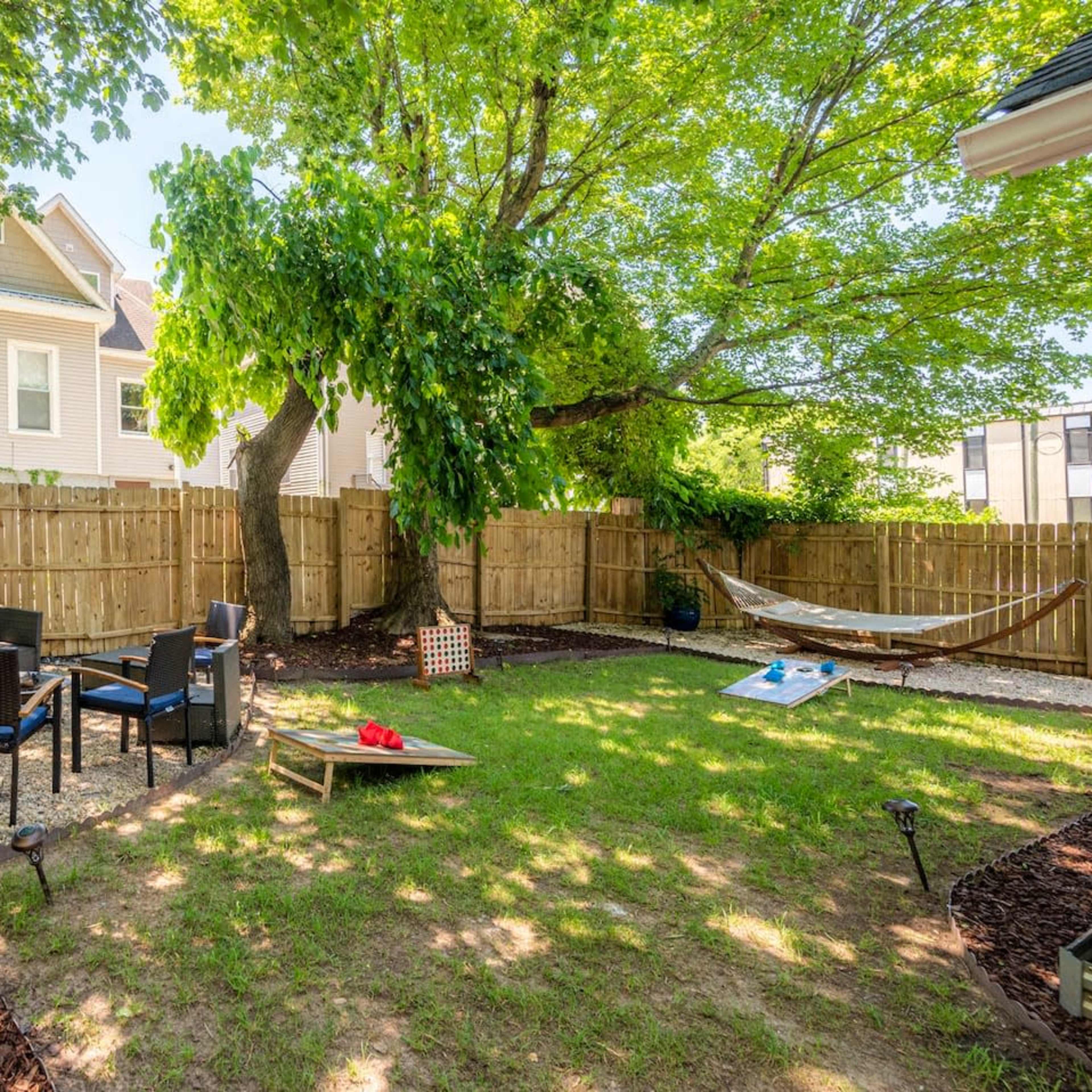 A backyard with a grassy area, a seating arrangement of chairs and a table, a hammock, and trees surrounded by a wooden fence.