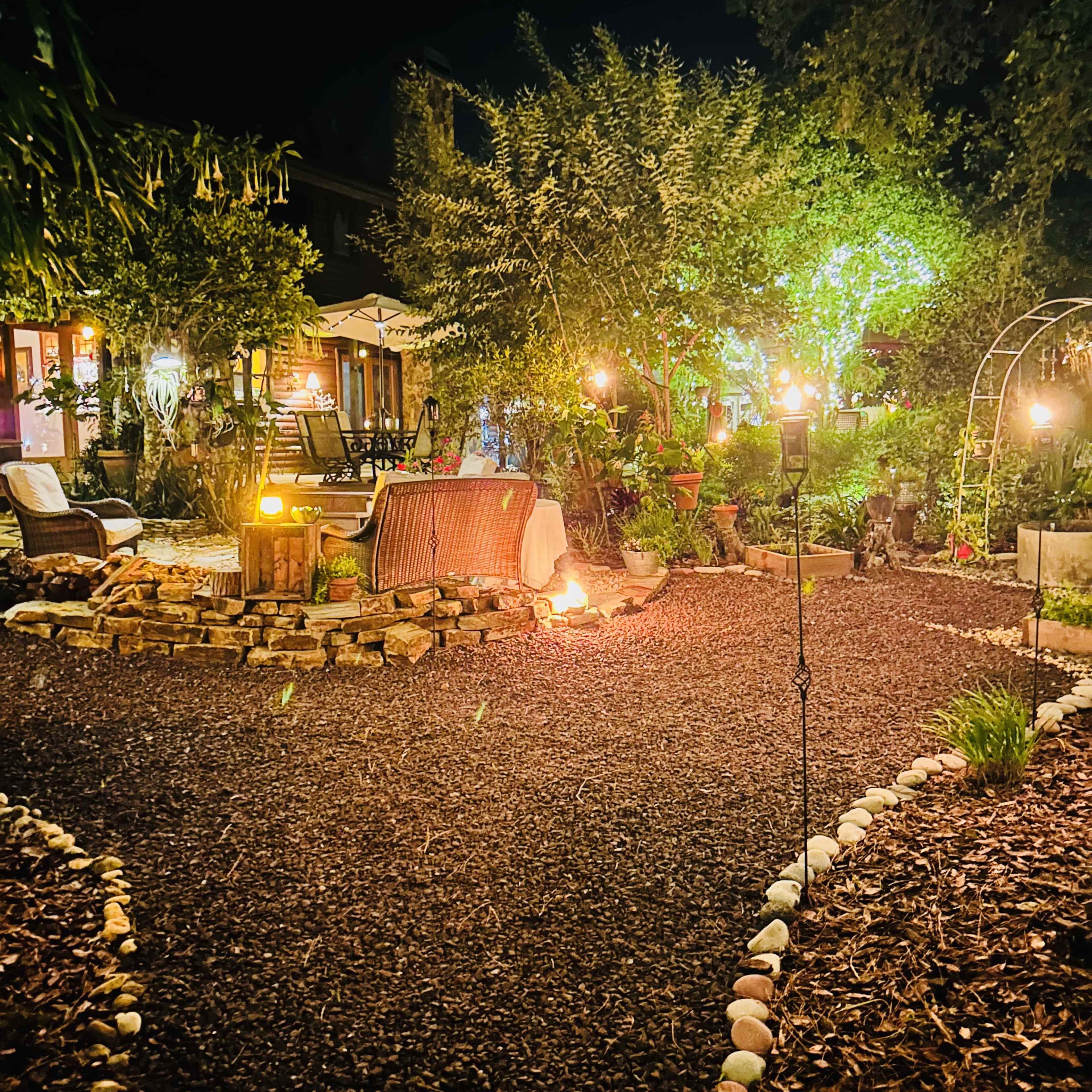 A pathway lined with stones leads through a gravel garden illuminated by soft lights, featuring seating areas and greenery.