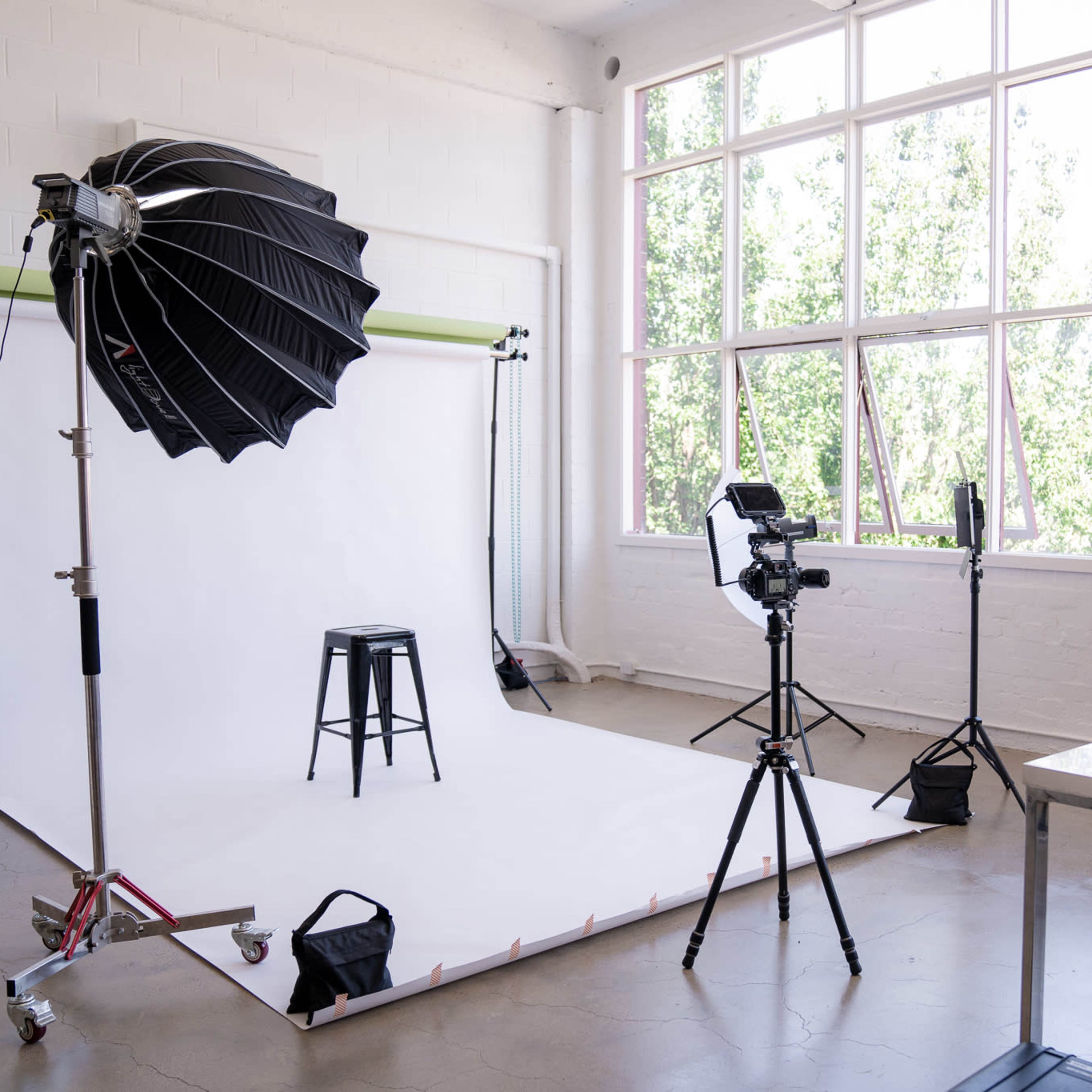 A photography studio setup with a white backdrop, a stool, various lighting equipment, and a camera on a tripod.