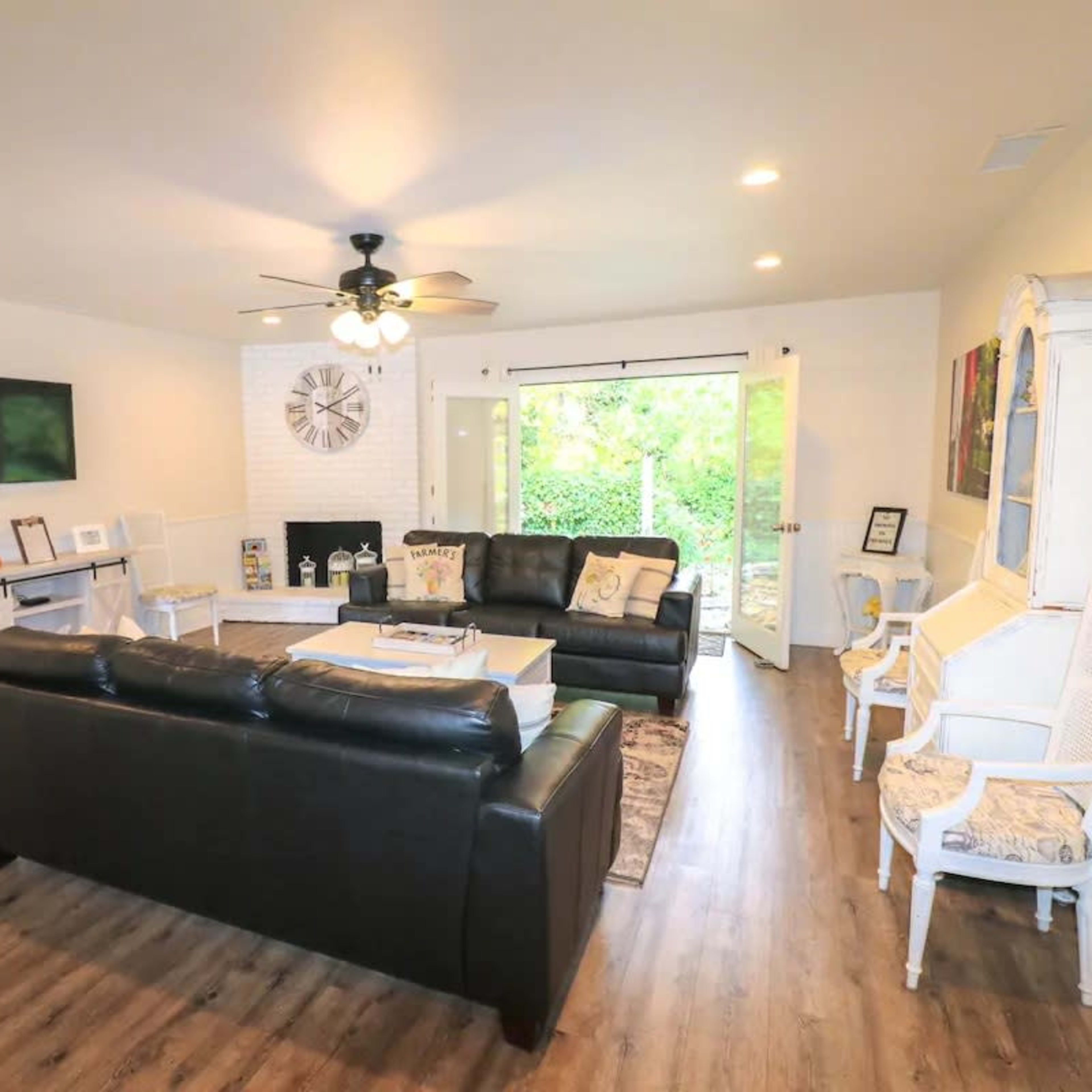 A spacious living room with a black leather sofa, a wall-mounted television, and large sliding doors leading to a green outdoor area.