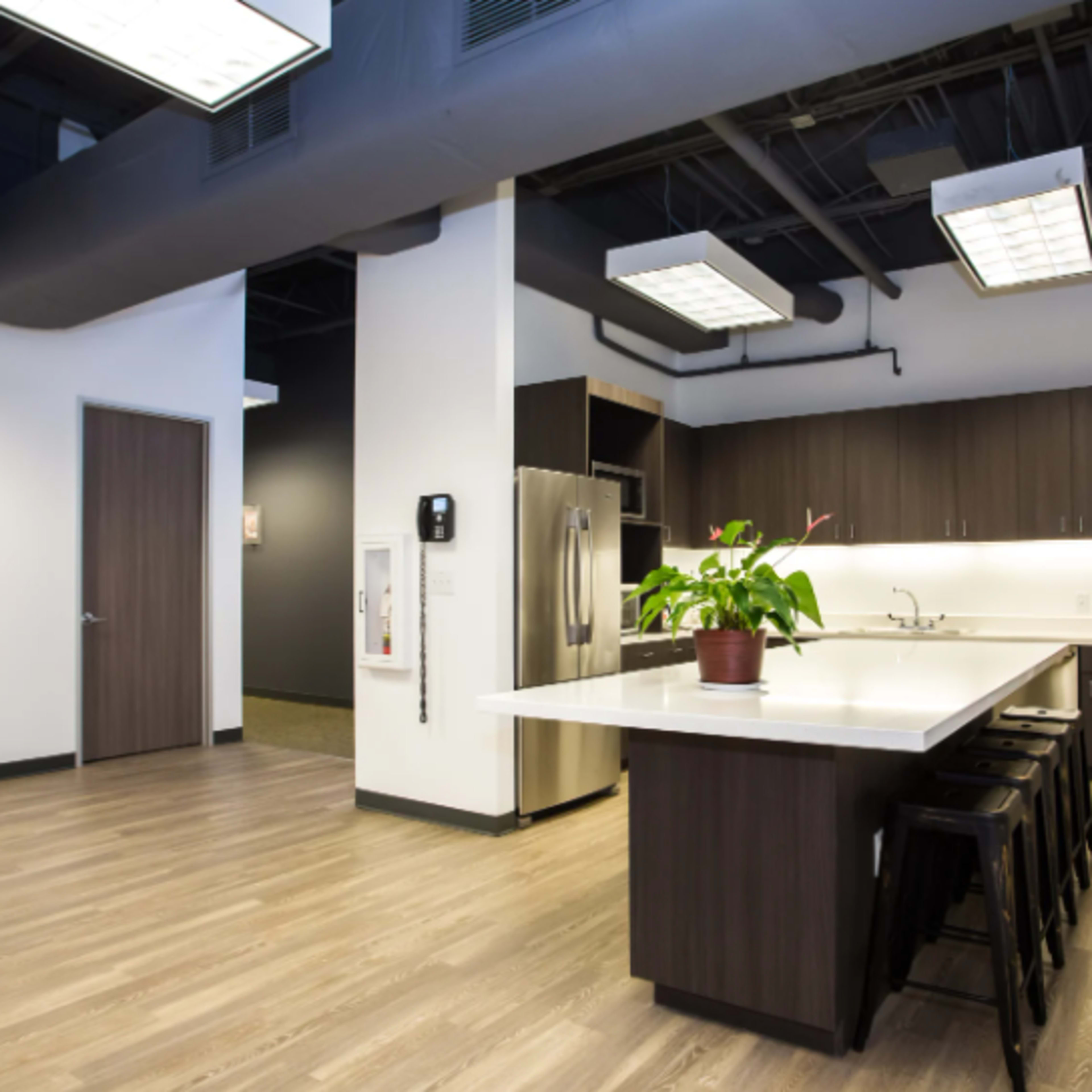 A modern kitchen area within an office, featuring a large island with seating, stainless steel appliances, and dark cabinetry.