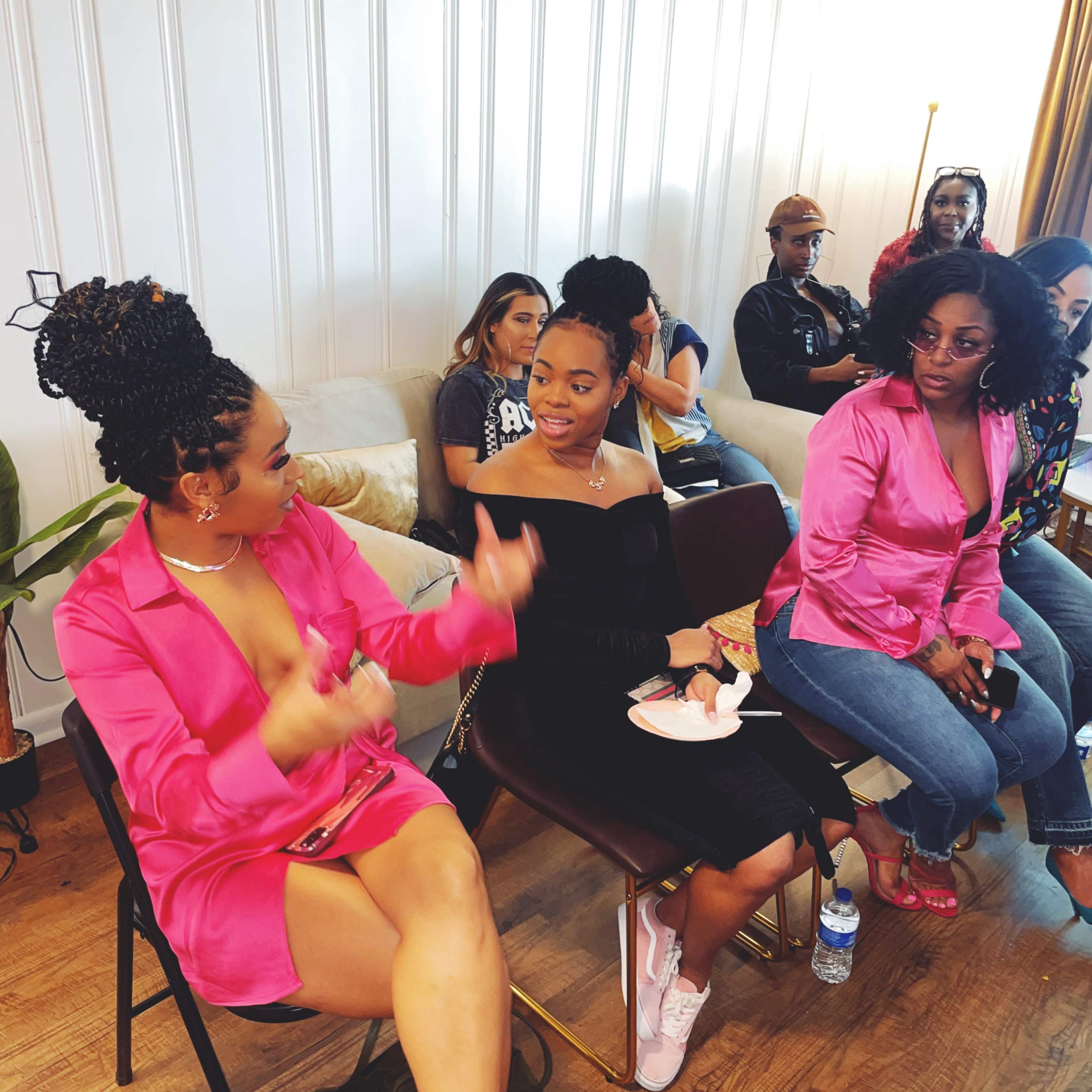A group of people sits in a modern, well-lit room, with two women engaged in conversation while others observe.