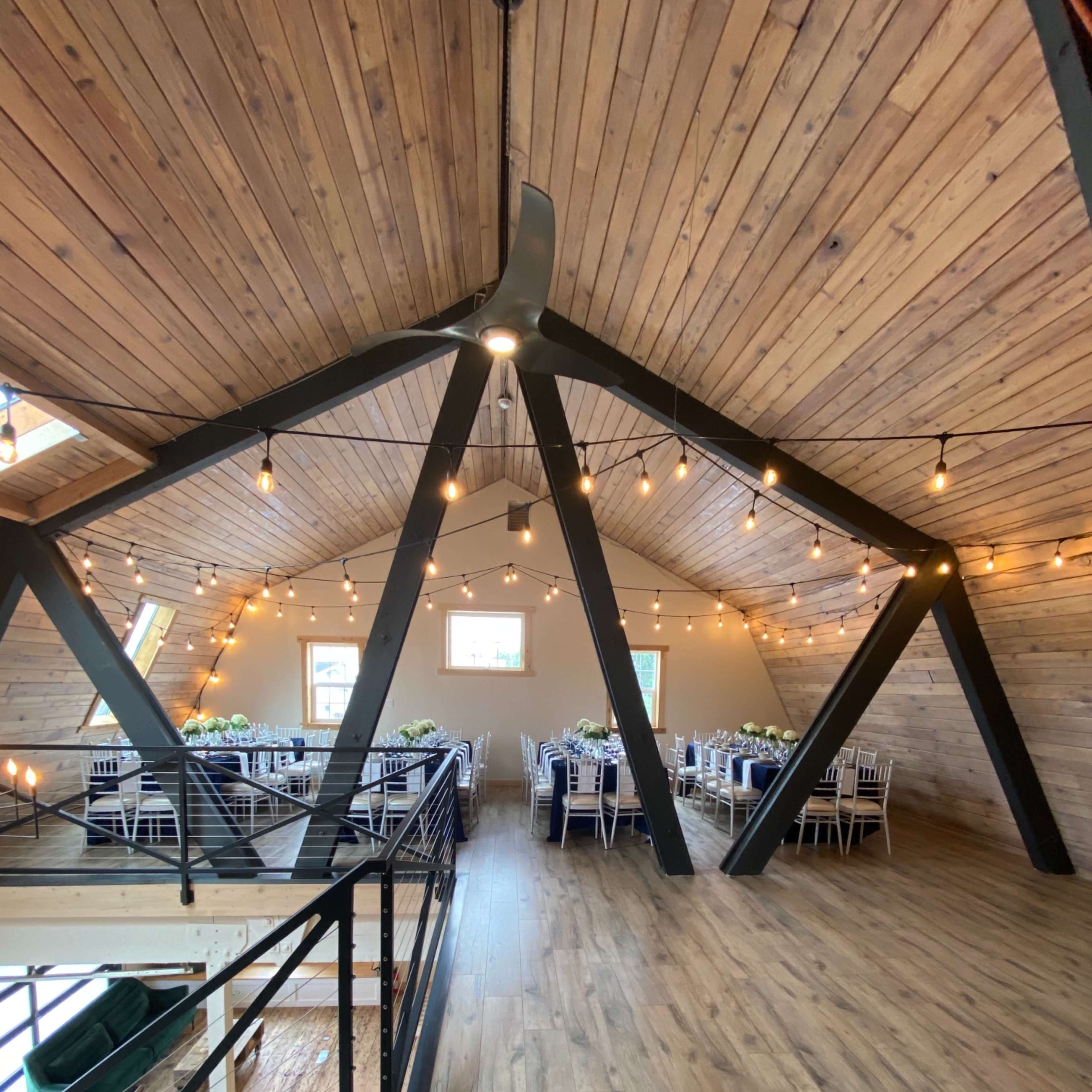 A spacious room with exposed wooden beams, a wooden floor, and tables set for dining beneath decorative string lights.