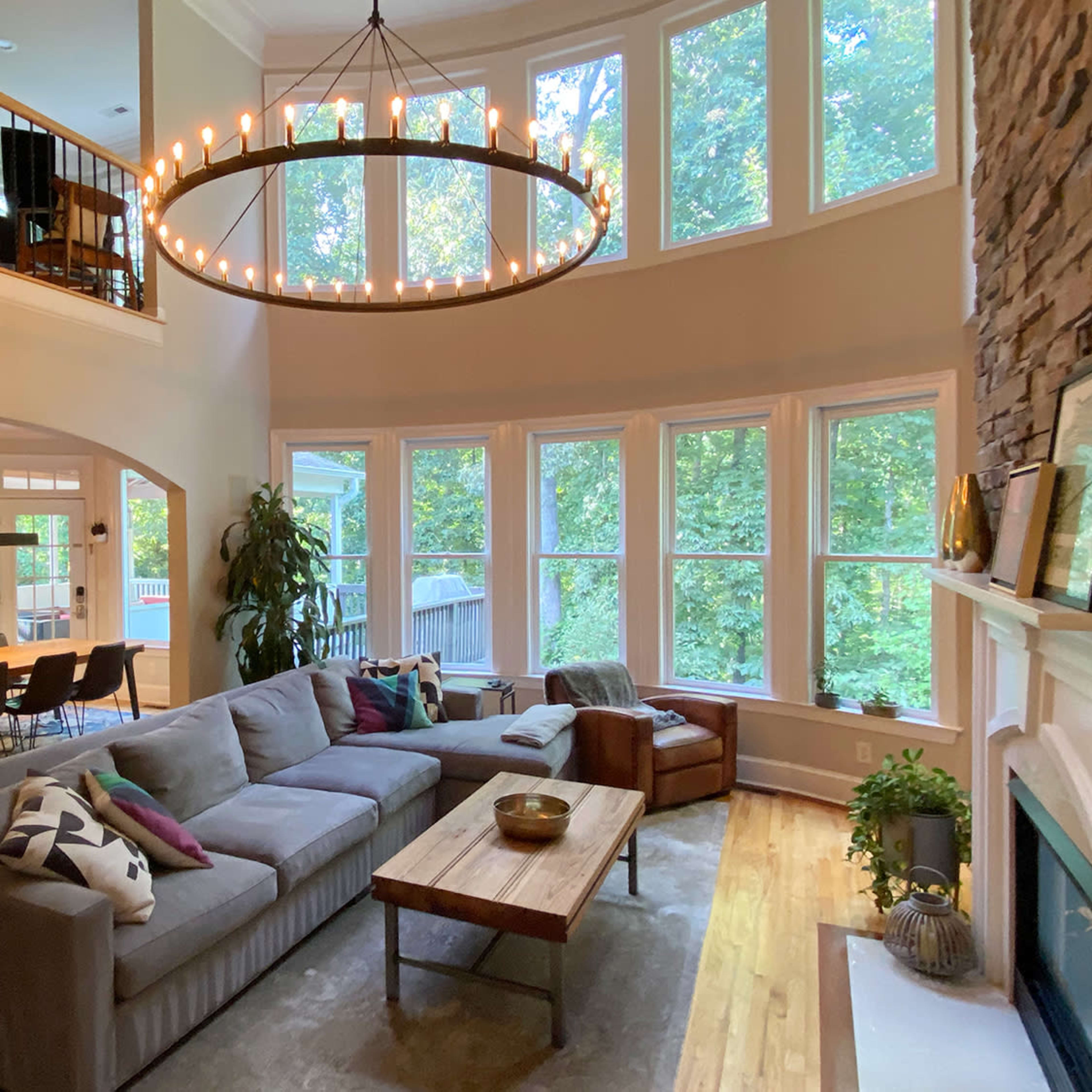 A spacious living room with large windows, a circular chandelier, a gray sectional sofa, a wooden coffee table, and a stone fireplace, overlooking a wooded area.