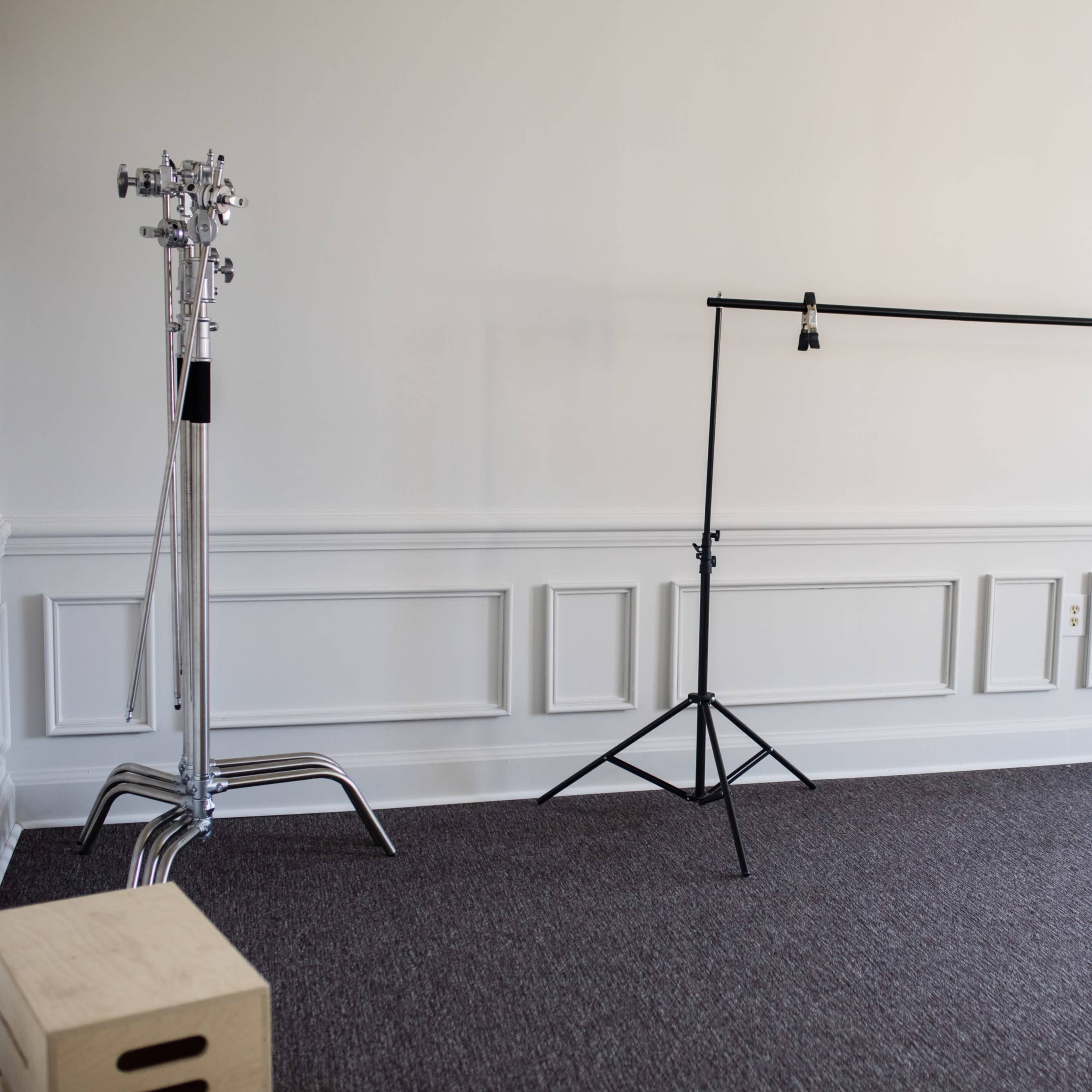 A minimalist room with a carpeted floor, featuring a black backdrop support frame and a metal light stand against a white paneled wall.