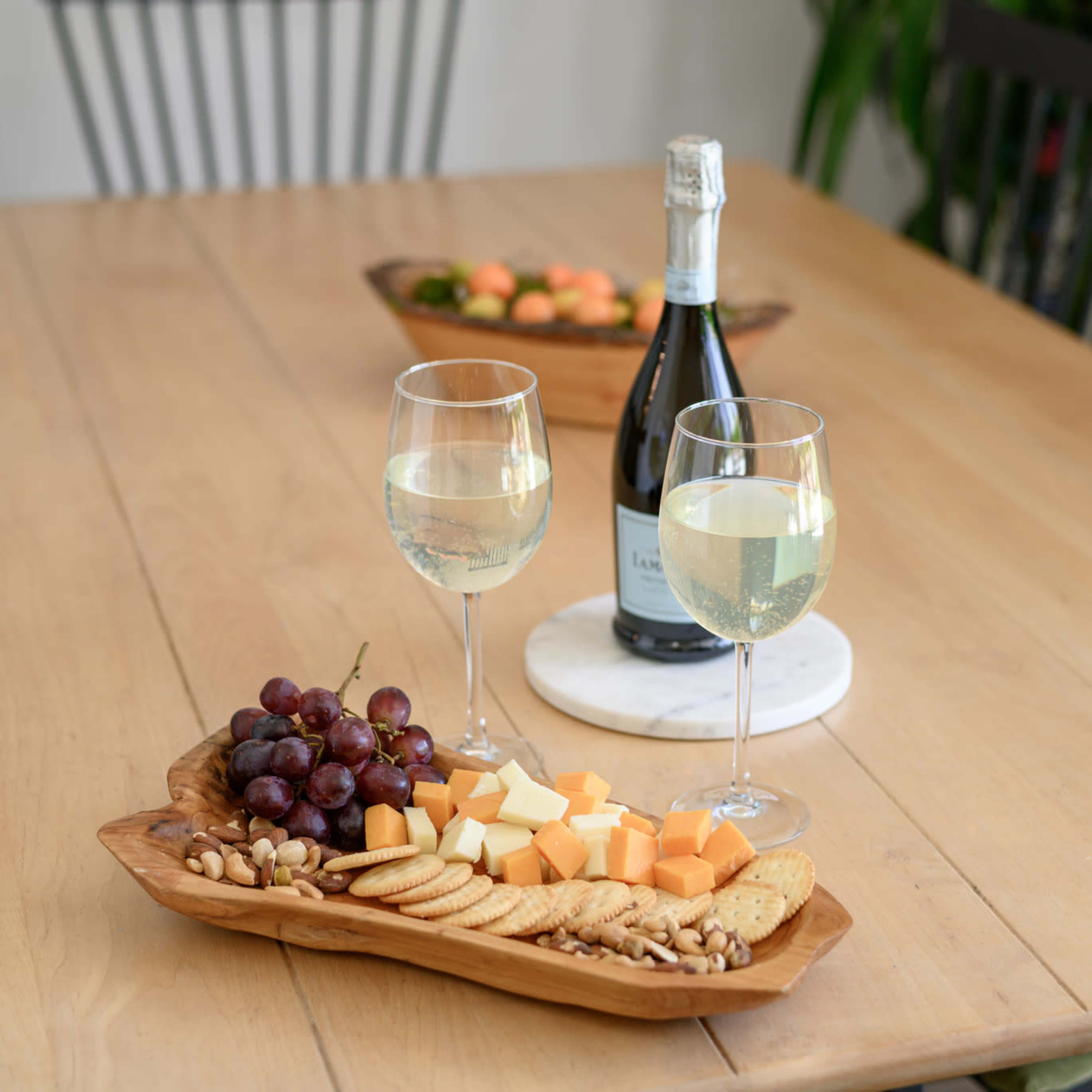 A wooden table displays a platter of assorted cheeses, grapes, and crackers alongside two glasses of sparkling wine and a bottle in the background.