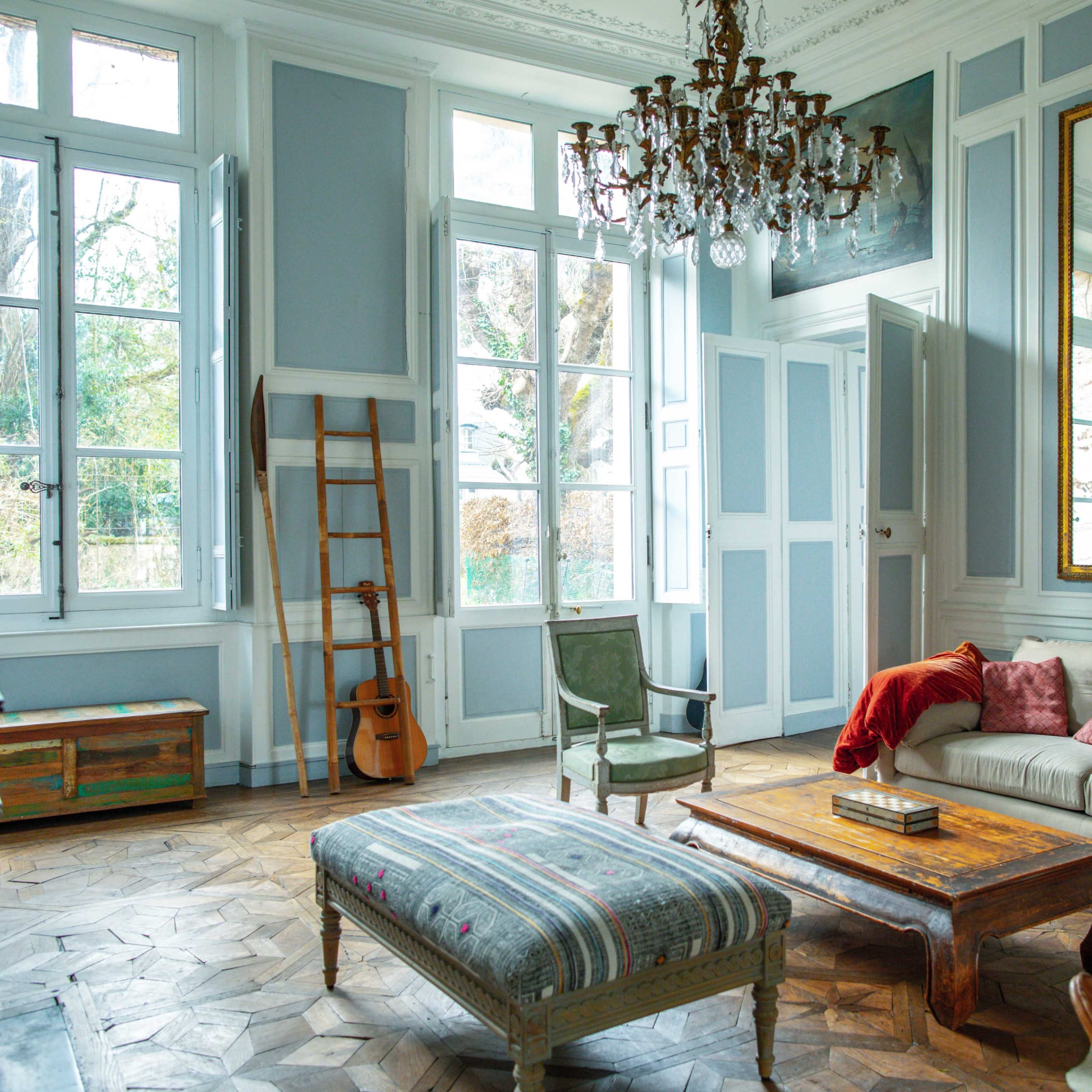 A spacious living room featuring a chandelier, large windows, a light-colored sofa with throw pillows, an ornate mirror, and wooden furniture.