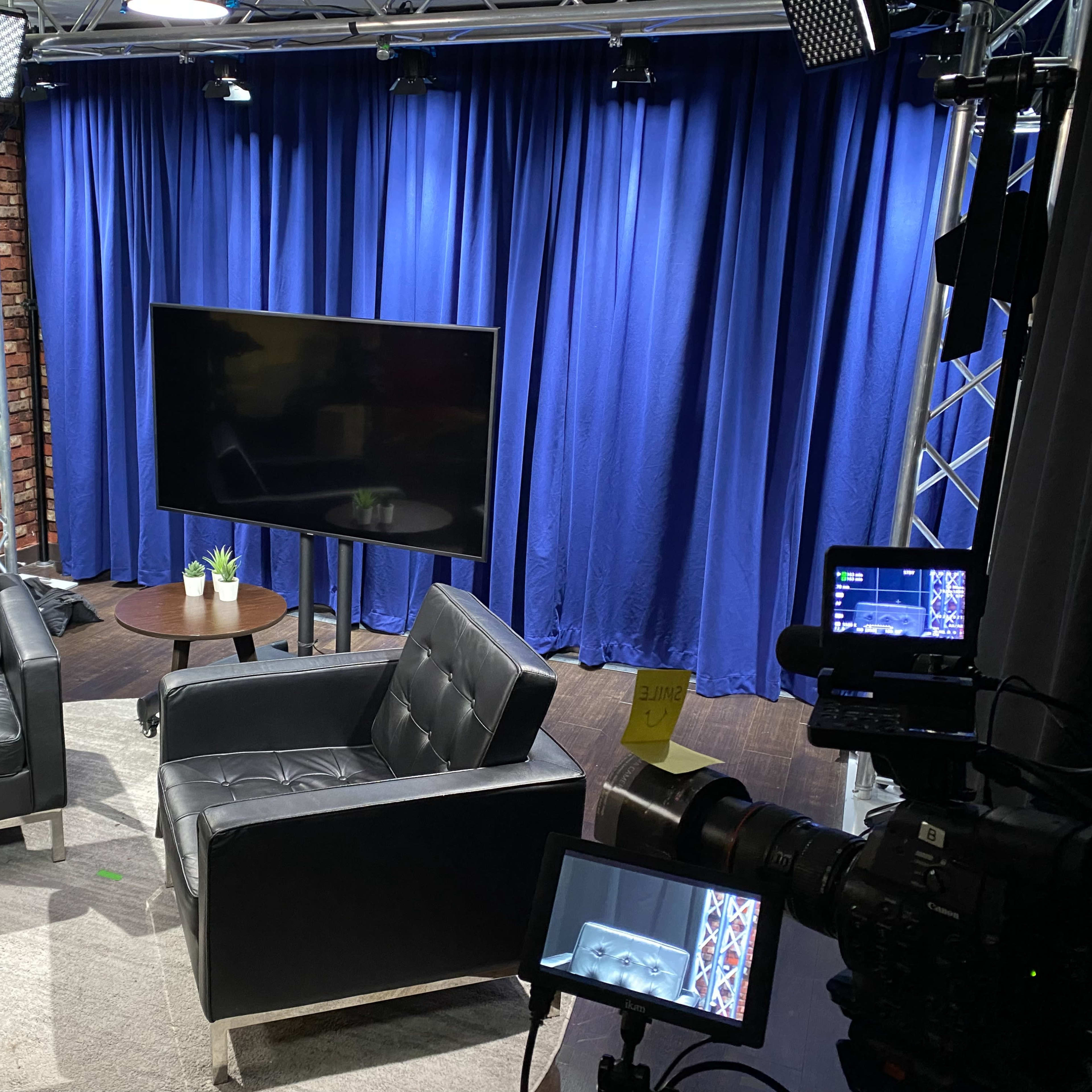 A television studio setup with two black chairs facing a monitor on a wooden table against a blue curtain backdrop.
