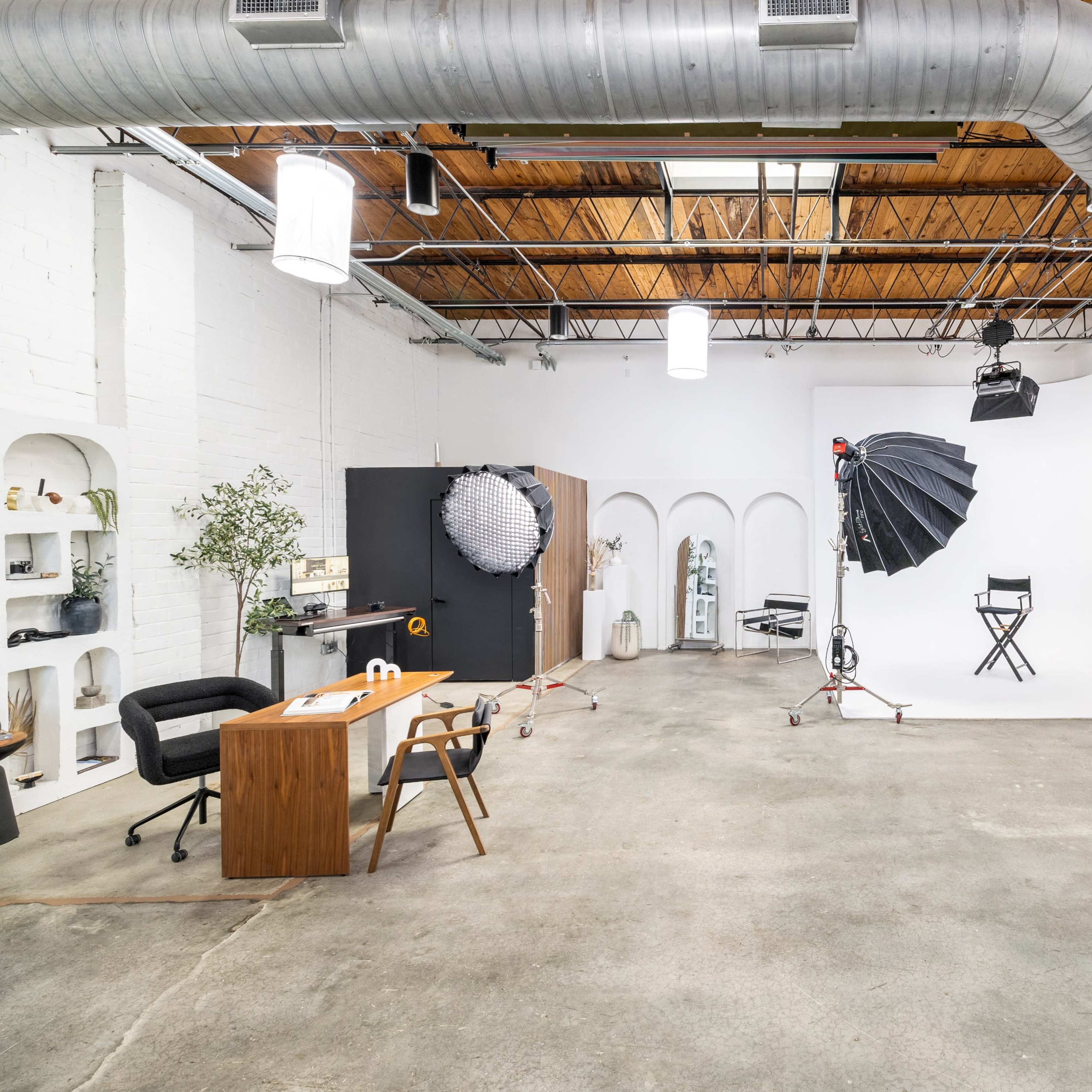 A spacious photography studio featuring various equipment, a desk with a chair, and a backdrop setup with lighting.