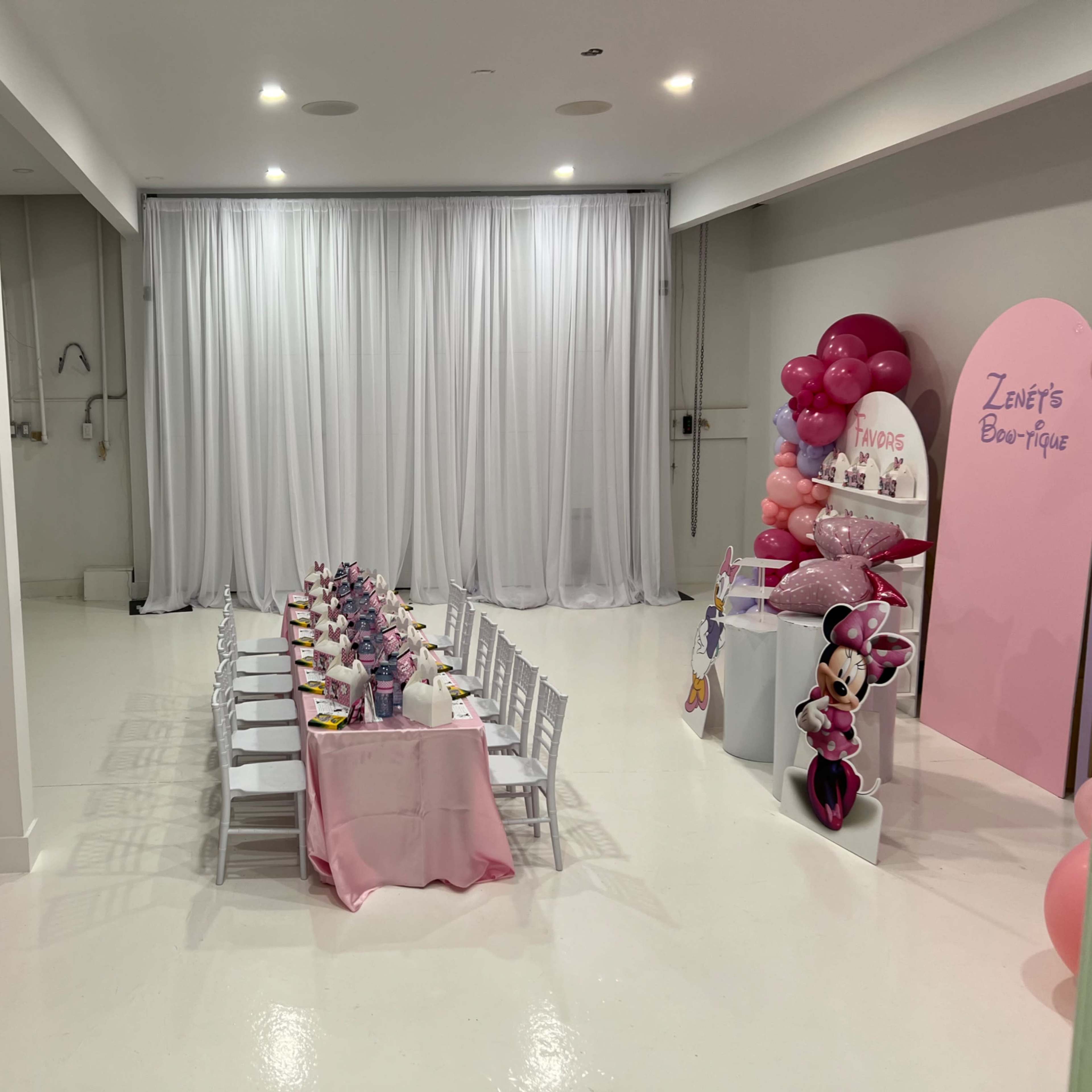 A decorated event space featuring a long dining table set with pink tableware and balloons, with white walls and a curtain backdrop.