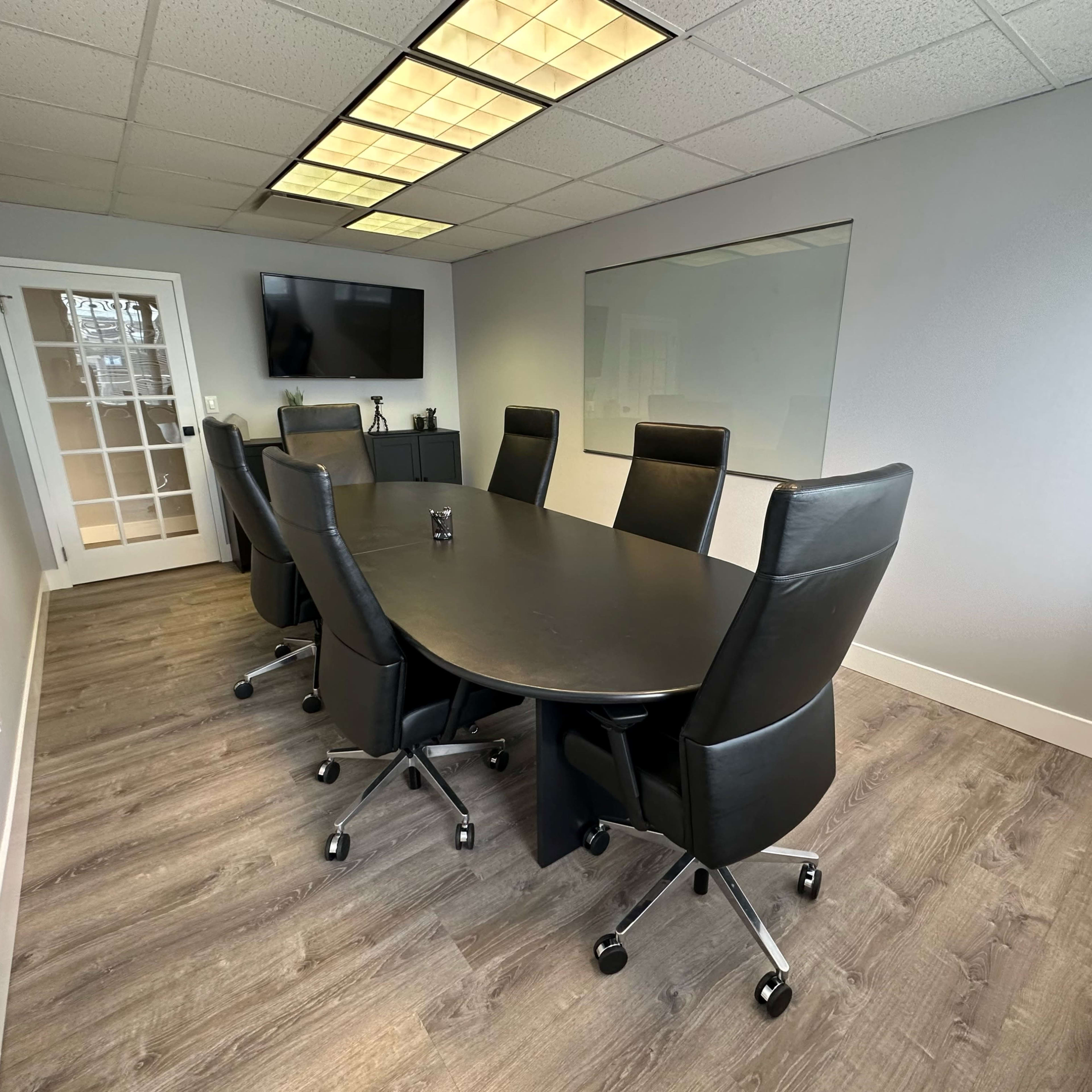 A modern conference room features a large oval table surrounded by black leather chairs, with a glass door and a wall-mounted screen in the background.