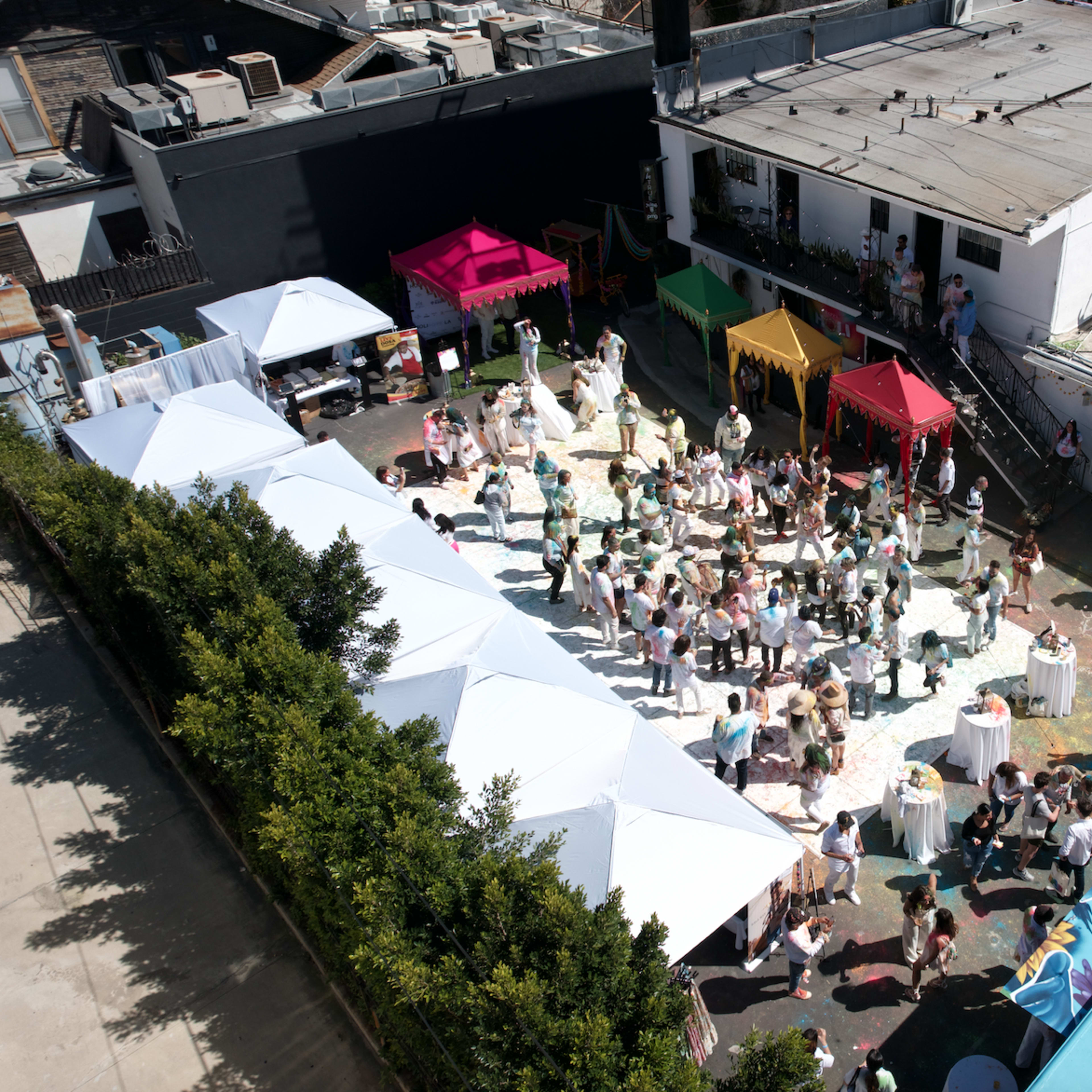 A large outdoor event with people dancing and socializing under colorful tents, surrounded by a white building and various food stands.