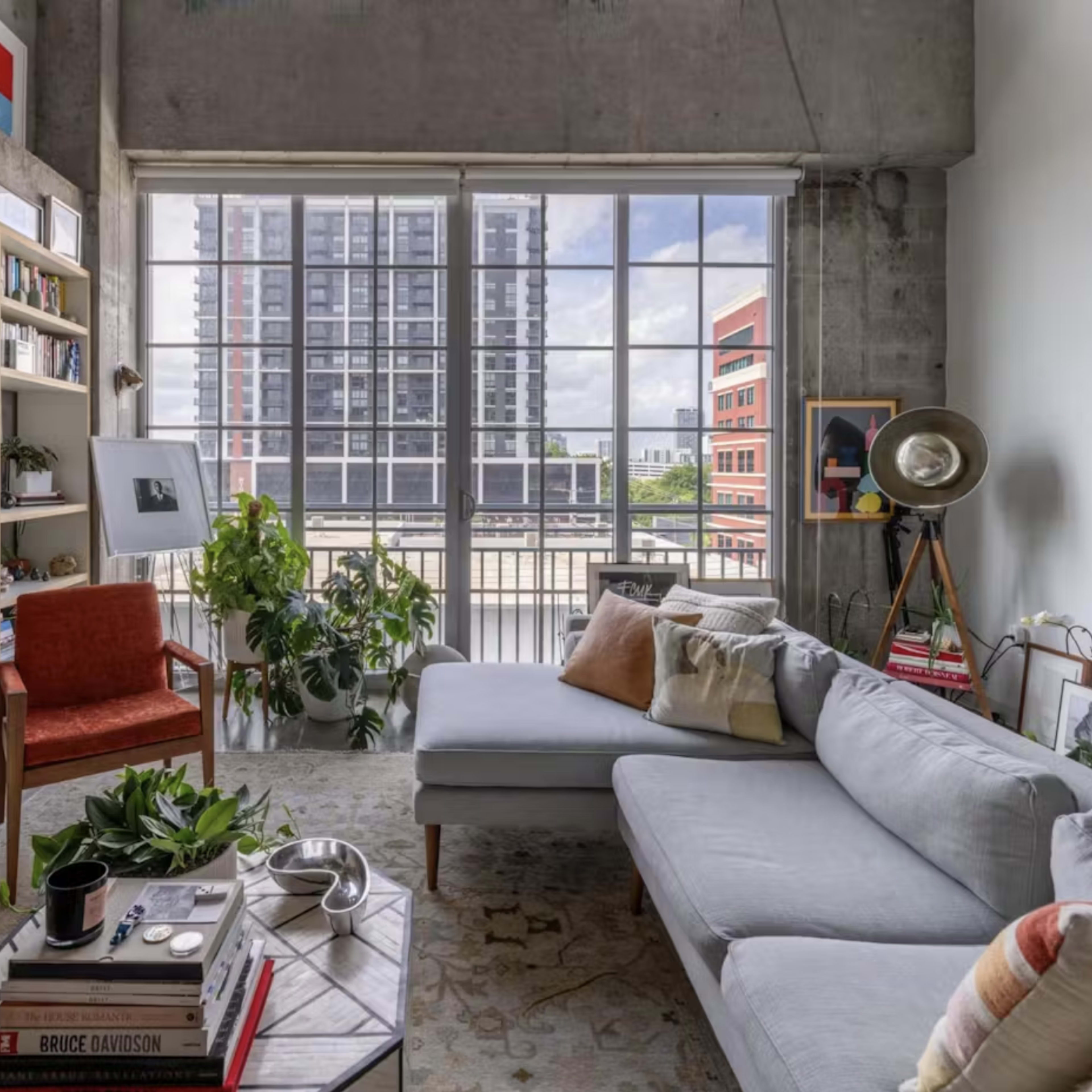 A modern living room features a light gray sectional sofa, shelves filled with books and decor, plants, and large windows showcasing a city view.