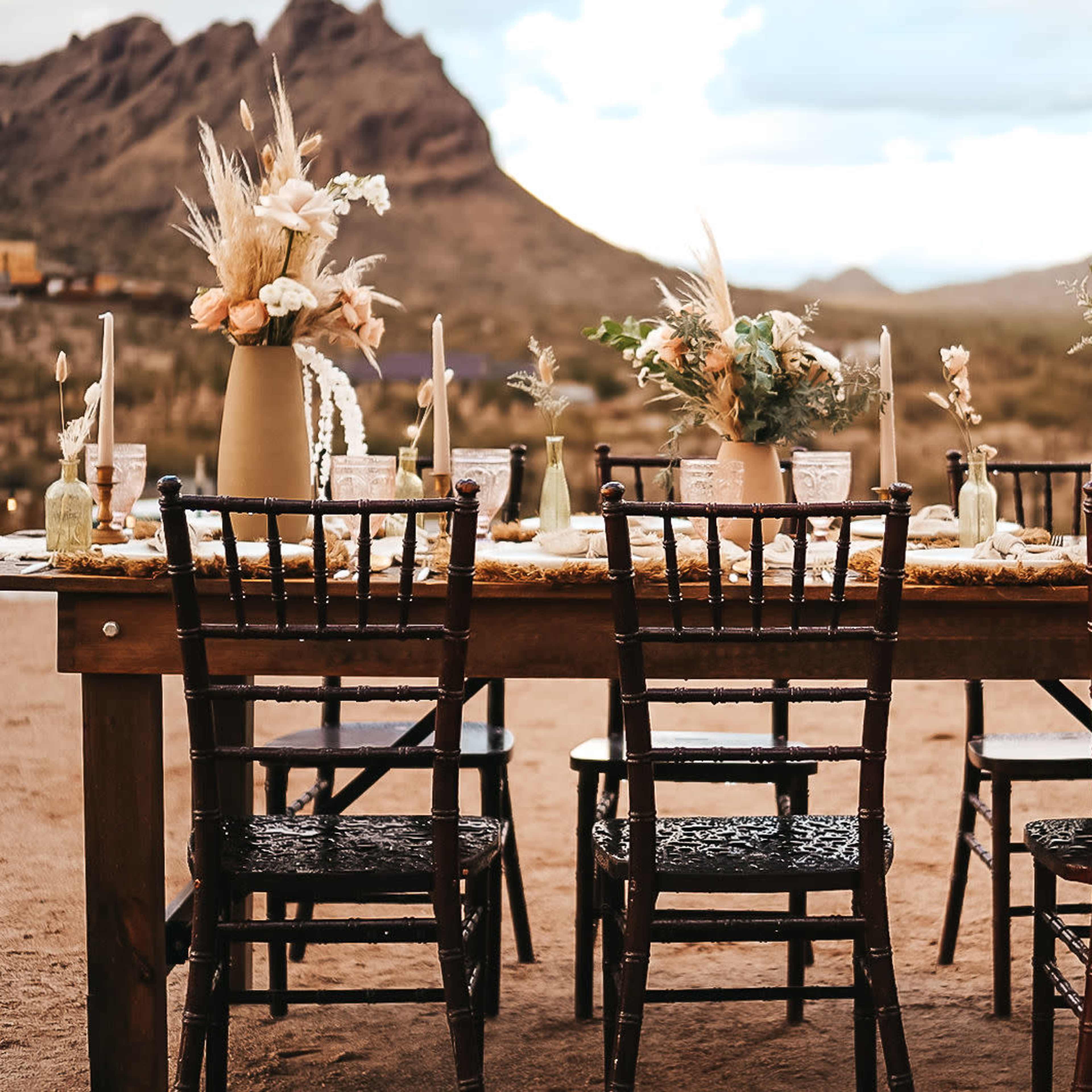 A rustic outdoor dining table is set with floral arrangements, candles, and chairs, surrounded by desert mountains.